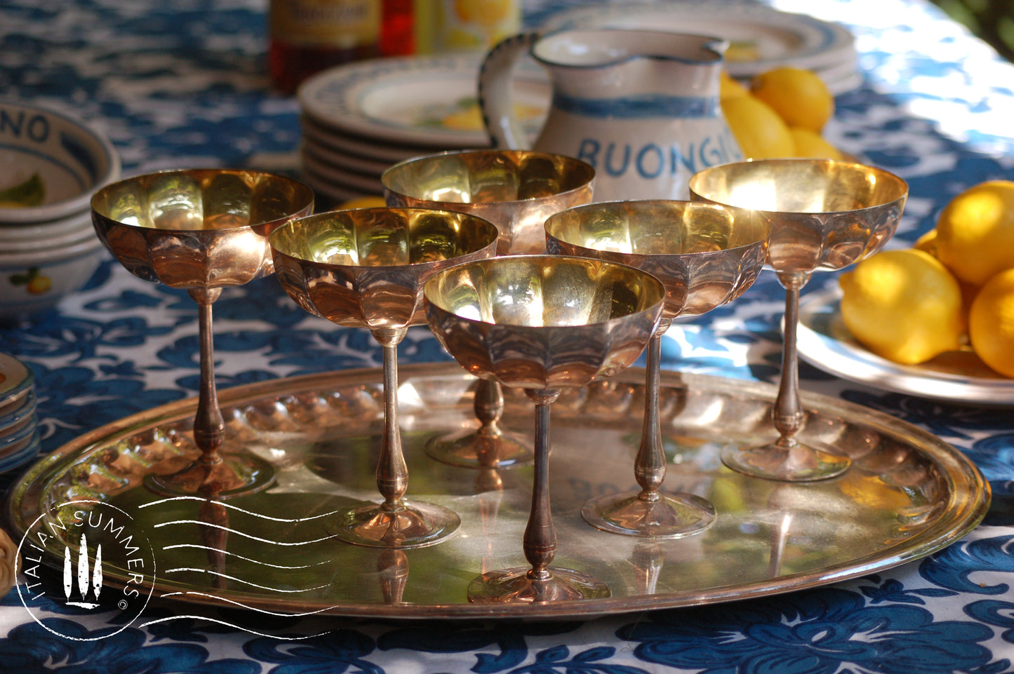 Italian Vintage Champagne coupes.set of six silver and gold plated long stemmed coupes on a silver plated tray upon a table with a flower blue Mediterranean pattern. A bowls with lemons is nearby along with white and blue maiolica serving bowls.,