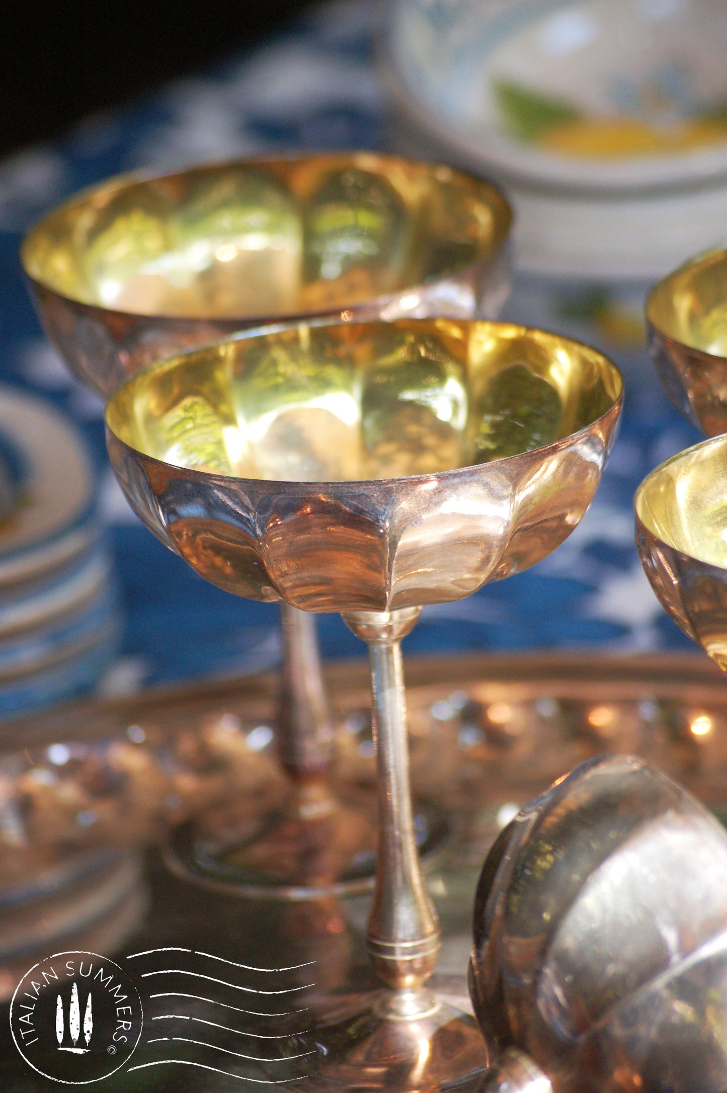 Italian Vintage Champagne coupes.set of six silver and gold plated long stemmed coupes on a silver plated tray upon a table with a flower blue Mediterranean pattern. A bowls with lemons is nearby along with white and blue maiolica serving bowls.,