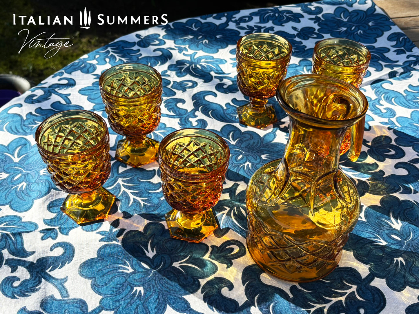 Set of Italian amber glass goblets and a carafe with the original Bormioli box on a floral patterned tablecloth.