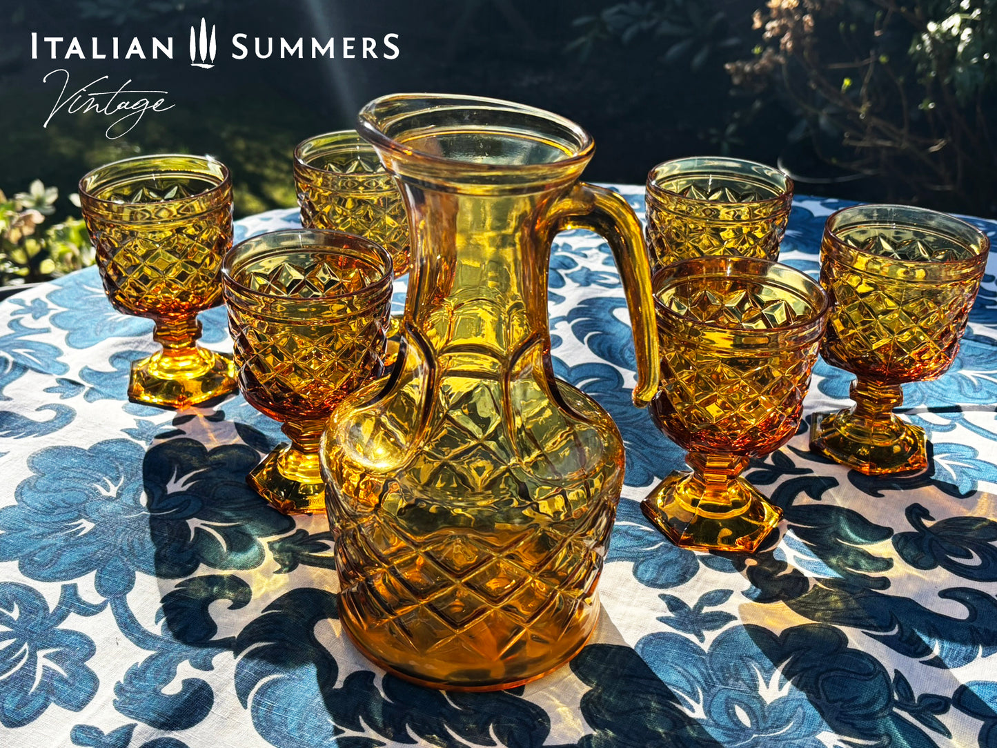 Set of Italian amber glass goblets and a carafe with the original Bormioli box on a floral patterned tablecloth.