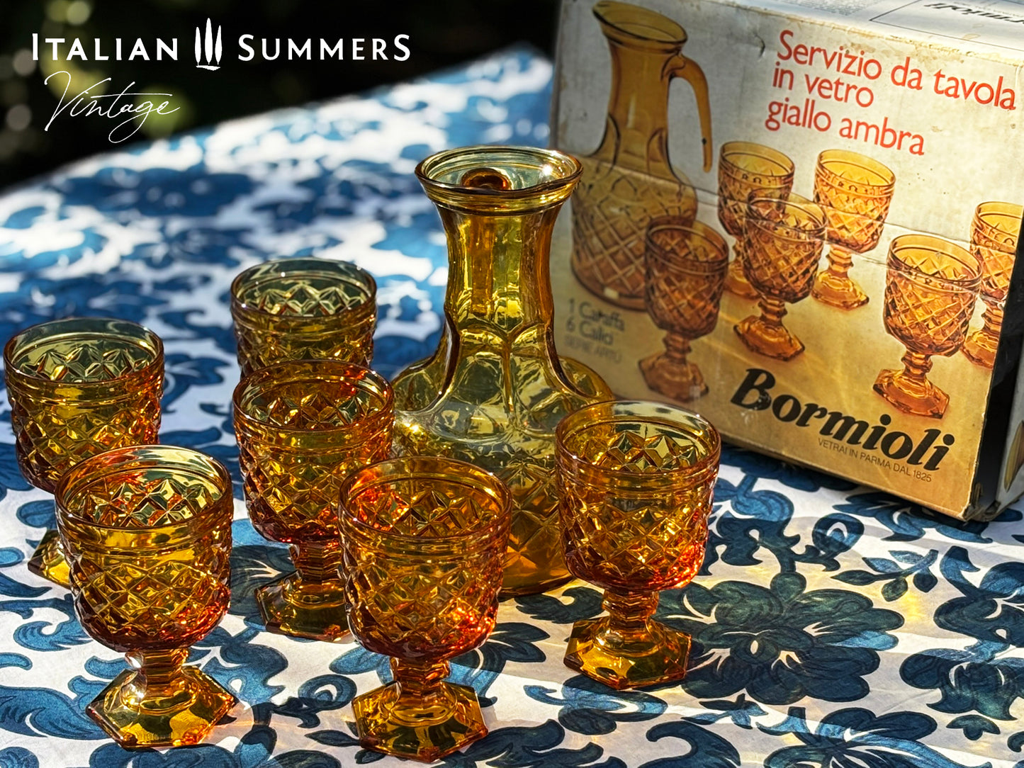Set of Italian amber glass goblets and a carafe with the original Bormioli box on a floral patterned tablecloth.