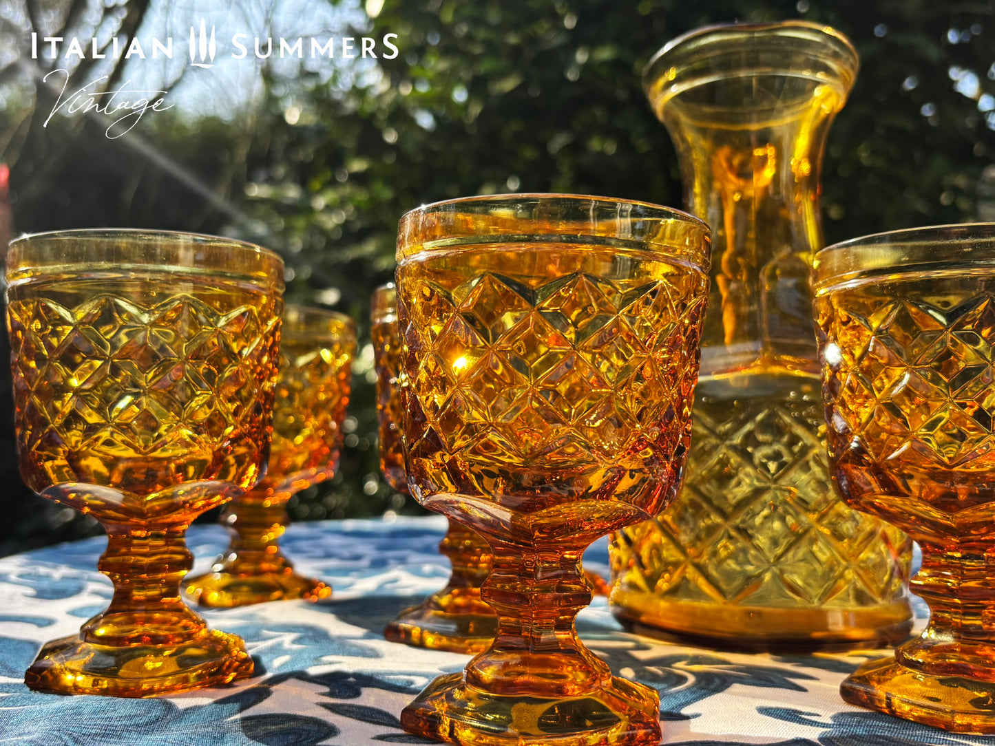 Set of Italian amber glass goblets and a carafe with the original Bormioli box on a floral patterned tablecloth.
