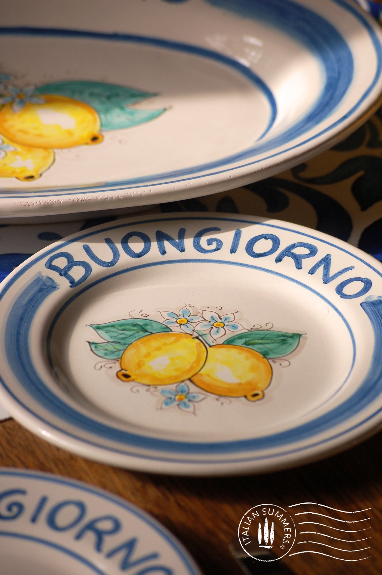 Ceramic plate Buongiorno Limoni handpainted in Sicily. This  plate has the quote Biongiorno on the rim - handpainted  - And colorfull lemons on the center of the plate  with lemon flowers. On the rim of the plate there are blue paint stripes. The blue color is ultra marine. This plate is designed and sold by Italian Summers.