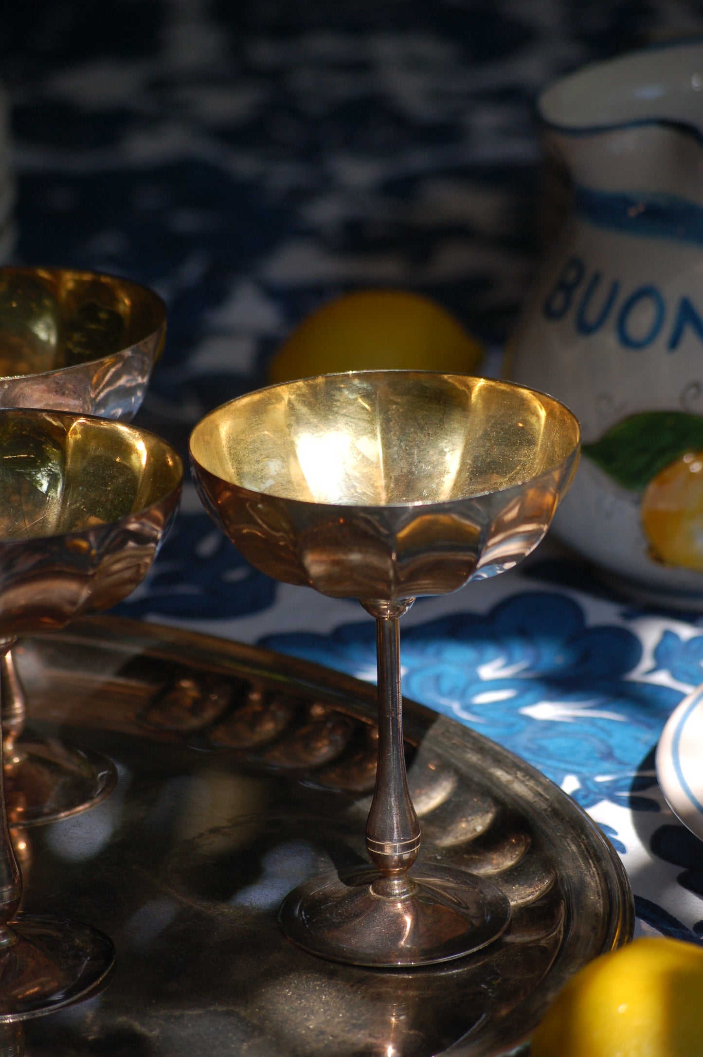 Italian Vintage Champagne coupes.set of six silver and gold plated long stemmed coupes on a silver plated tray upon a table with a flower blue Mediterranean pattern. A bowls with lemons is nearby along with white and blue maiolica serving bowls.,