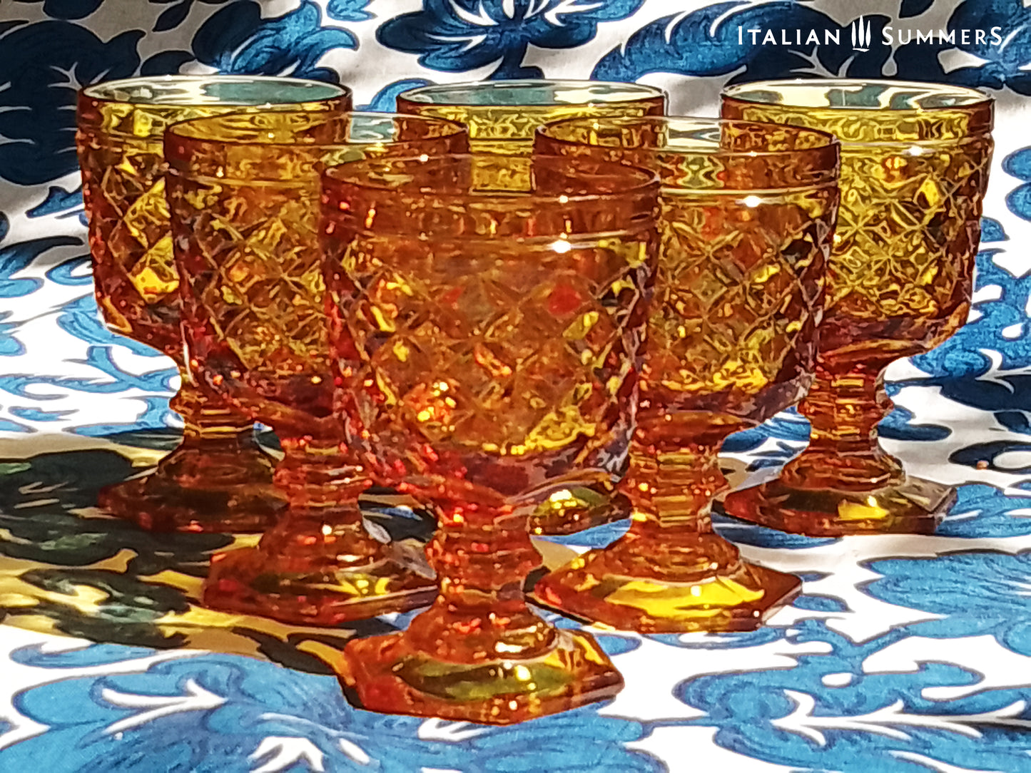 Set of Italian amber glass goblets and a carafe with the original Bormioli box on a floral patterned tablecloth.