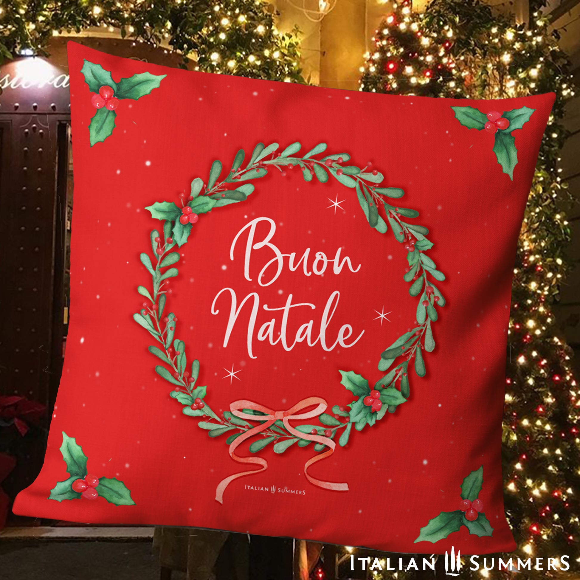 Red pillow with Italian Christmas design and text in front of a festive Italian village street scene.