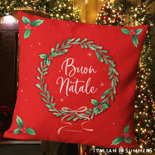 Red pillow with Italian Christmas design and text in front of a festive Italian village street scene.