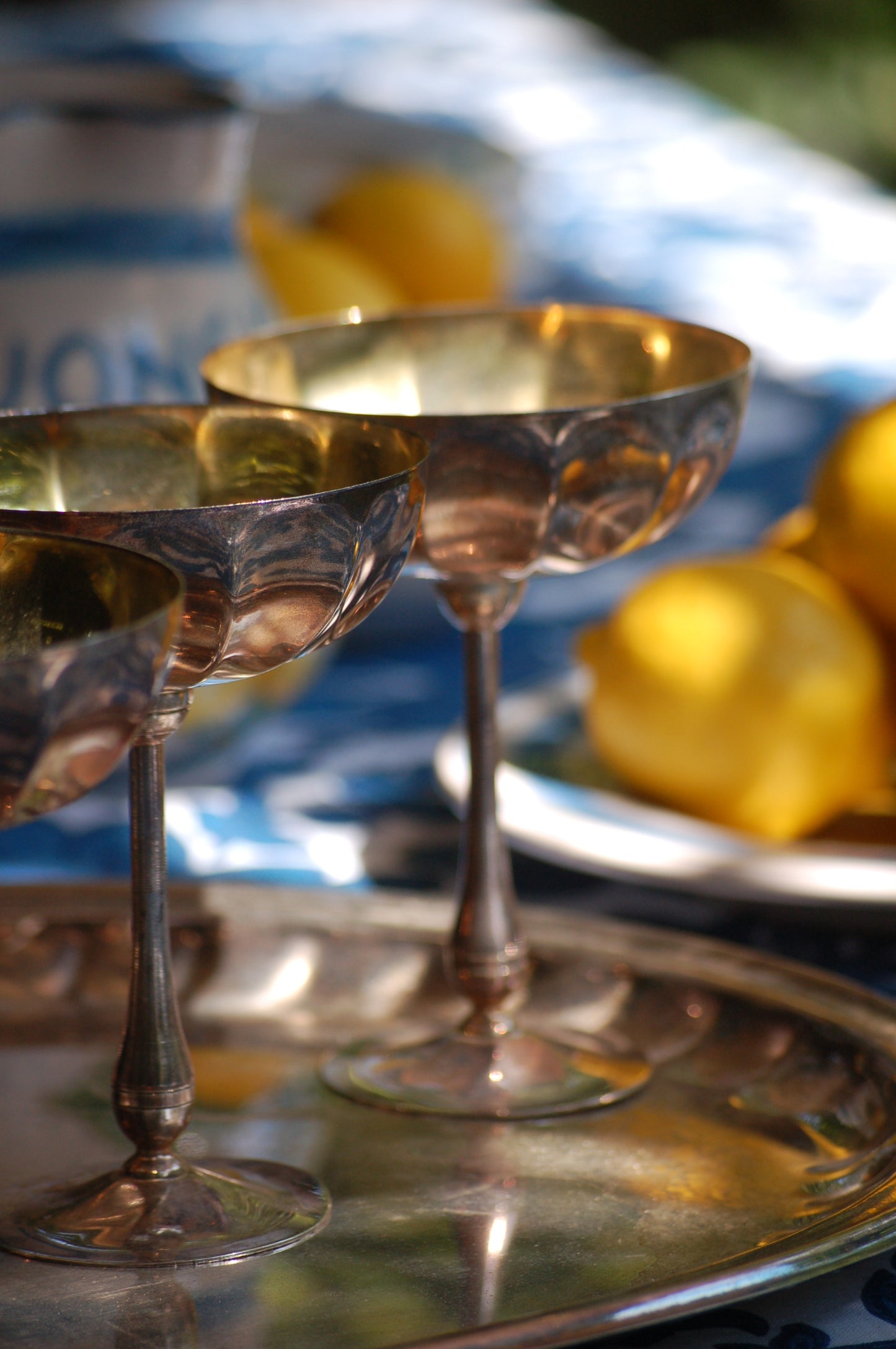 Italian Vintage Champagne coupes.set of six silver and gold plated long stemmed coupes on a silver plated tray upon a table with a flower blue Mediterranean pattern. A bowls with lemons is nearby along with white and blue maiolica serving bowls.,