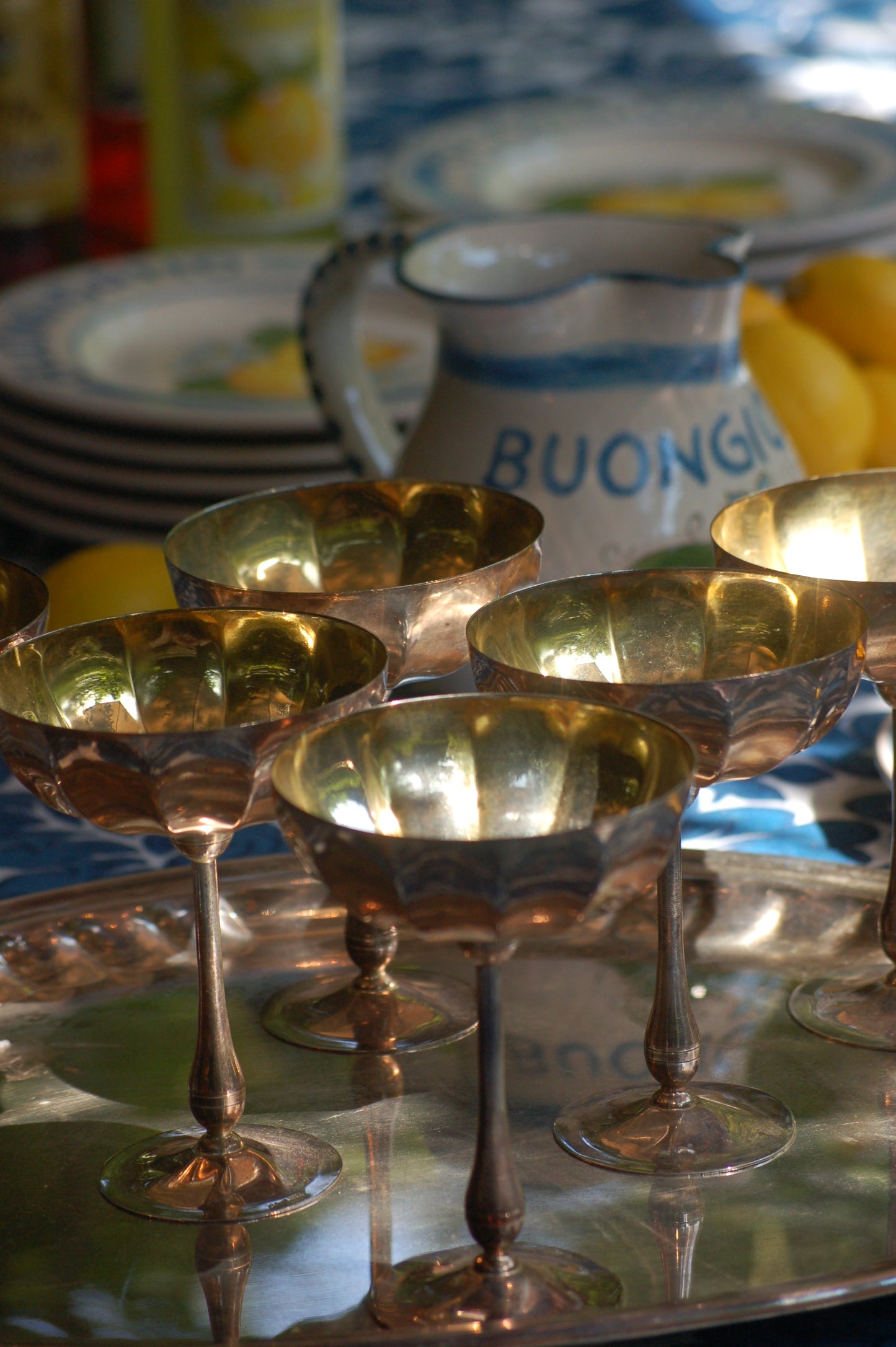 Italian Vintage Champagne coupes.set of six silver and gold plated long stemmed coupes on a silver plated tray upon a table with a flower blue Mediterranean pattern. A bowls with lemons is nearby along with white and blue maiolica serving bowls.,