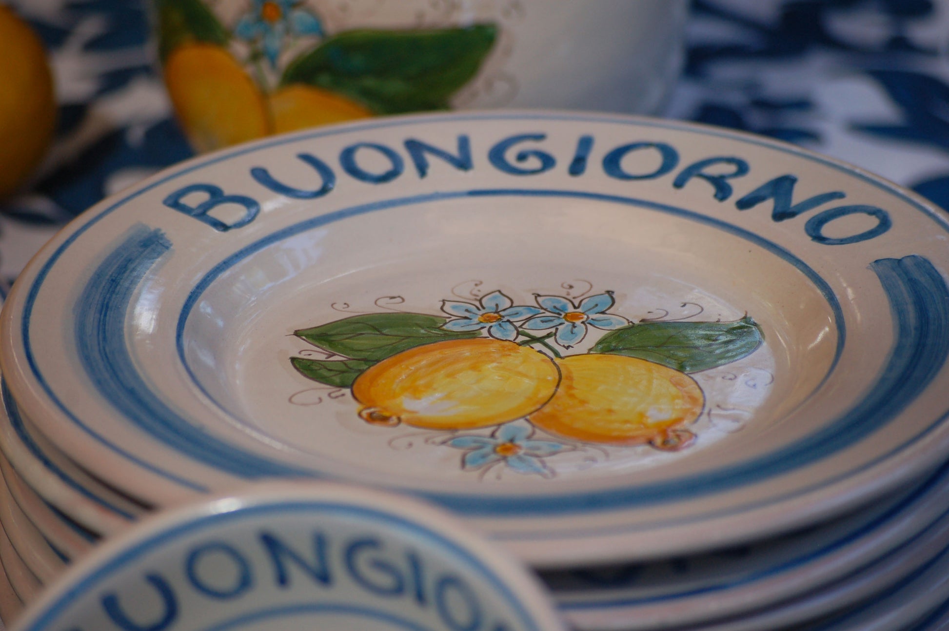 Ceramic plate Buongiorno Limoni handpainted in Sicily. This  plate has the quote Biongiorno on the rim - handpainted  - And colorfull lemons on the center of the plate  with lemon flowers. On the rim of the plate there are blue paint stripes. The blue color is ultra marine. This plate is designed and sold by Italian Summers.