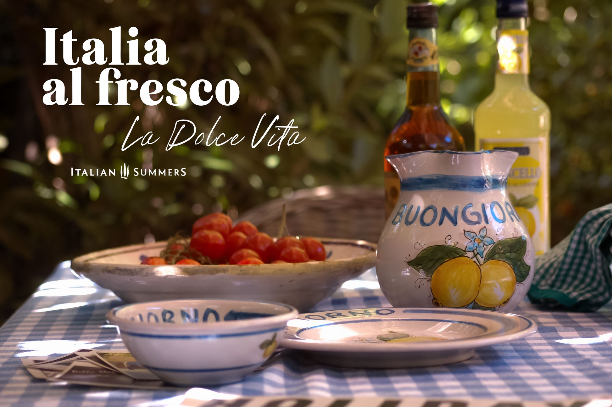 An Italy inspired table in summertime. Ont the table are Italian ceramics with lemons and the word Buongiorno. The is a plate, a bowl and a Buongiorno carafe. By Italian Summers