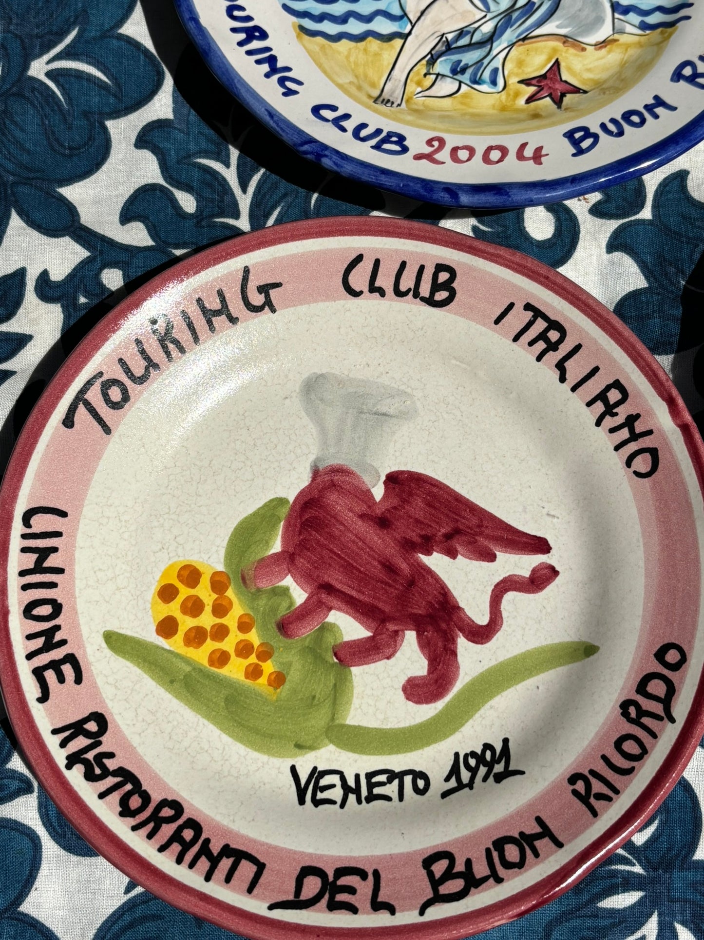 Italian vintage ceramic plate with a red lion of Venice (San Marco) and chef's hat design, text Touring Club Italiano on a blue floral tablecloth