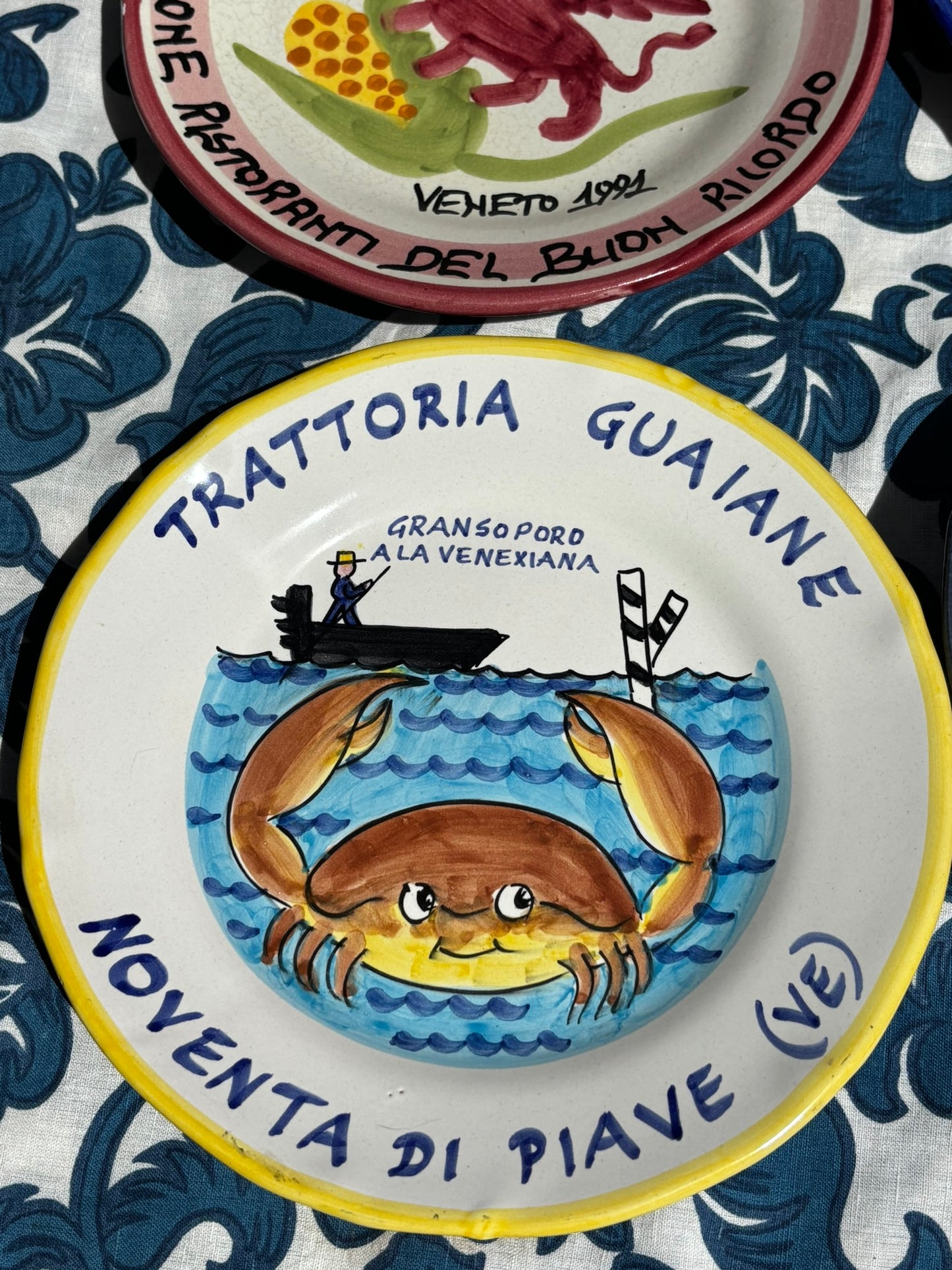 Italian ceramic plate with a crab illustration on the sea and Italian text Trattoria Guaiane Venezia on a blue and white patterned tablecloth