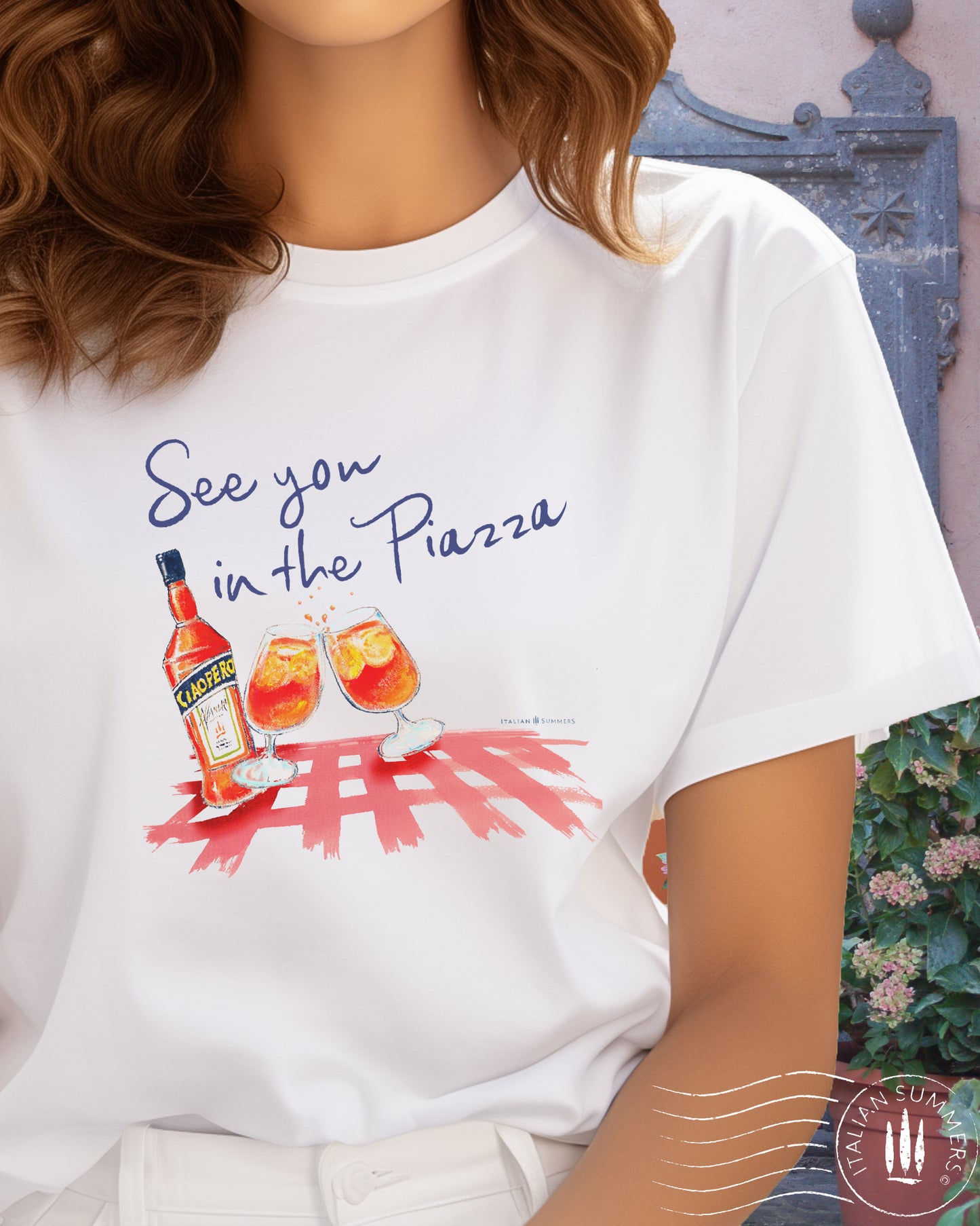 A girl wears an Italy inspired white t-shirt with the text See you in the Piazza in navy blue and a sketch in water color with an Aperol bottle and 2 glasses filled with Aperol Spritz on a bright red table cloth. Designed and sold by Italian Summers