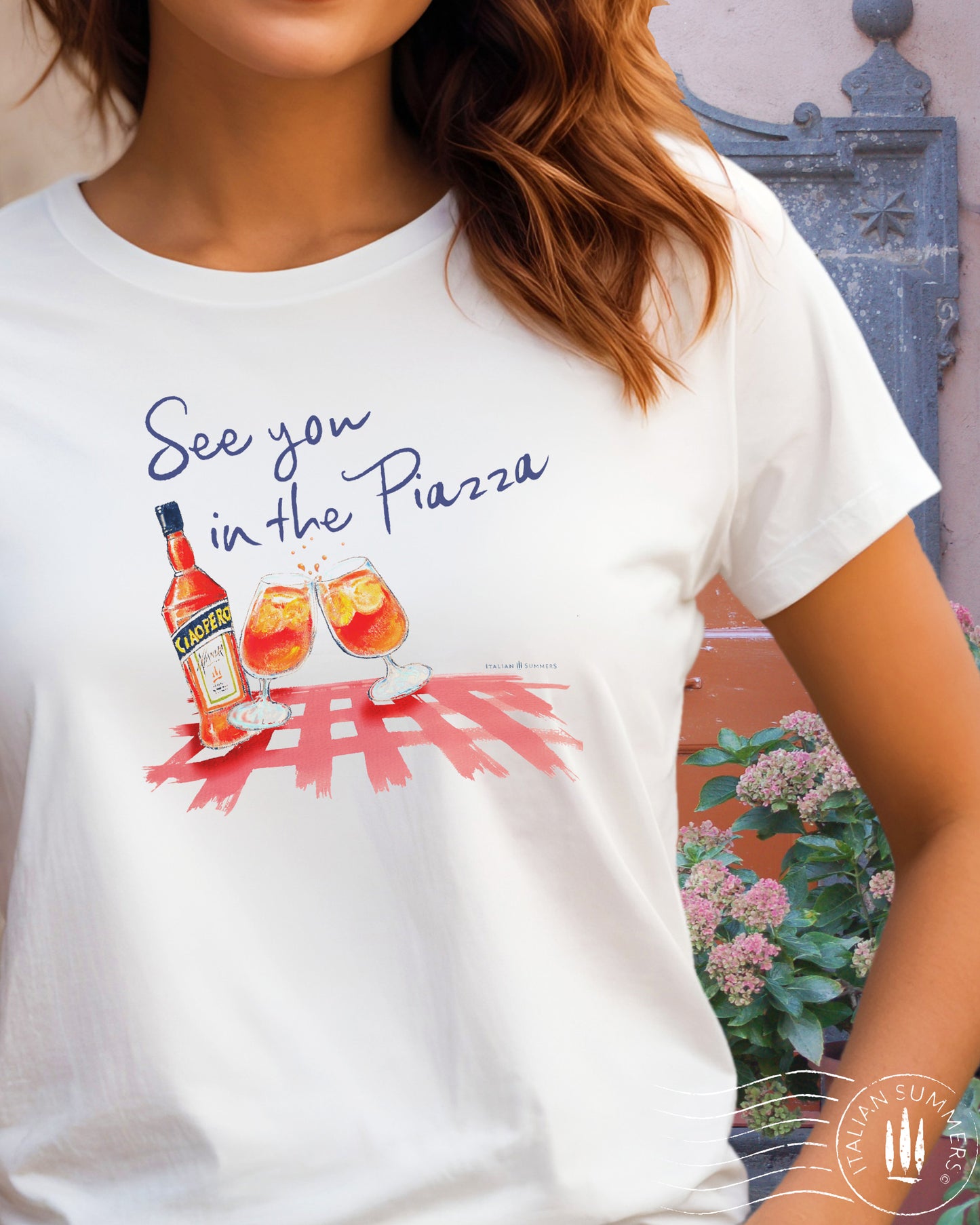 A girl wears an Italy inspired white t-shirt with the text See you in the Piazza in navy blue and a sketch in water color with an Aperol bottle and 2 glasses filled with Aperol Spritz on a bright red table cloth. Designed and sold by Italian Summers