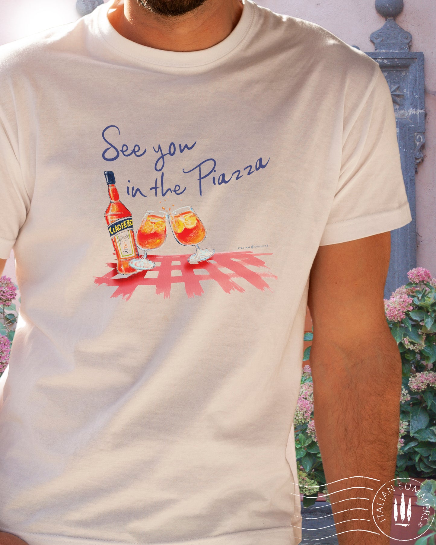 A girl wears an Italy inspired white t-shirt with the text See you in the Piazza in navy blue and a sketch in water color with an Aperol bottle and 2 glasses filled with Aperol Spritz on a bright red table cloth. Designed and sold by Italian Summers