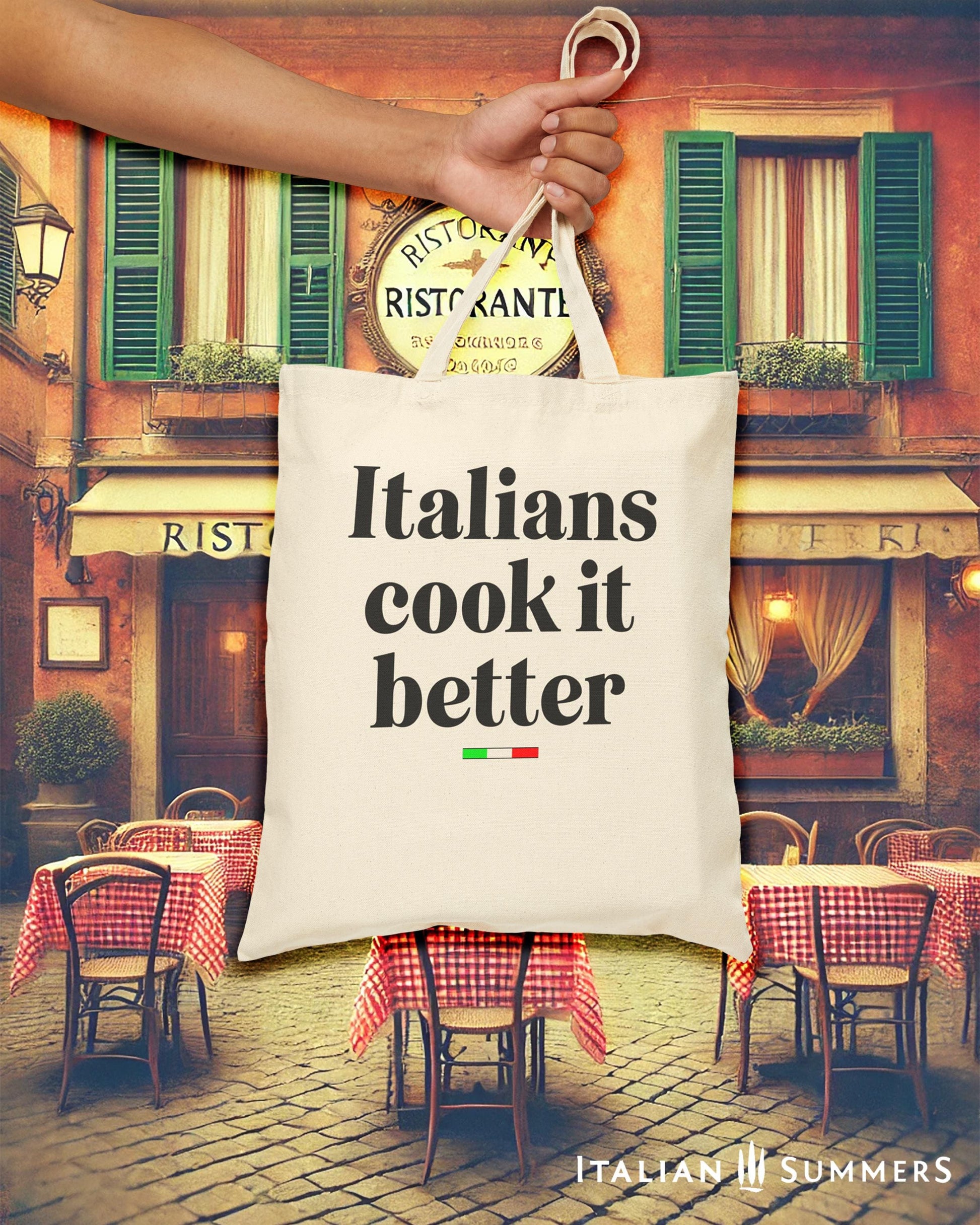 A Canvas tote bag with the quote Italians cook it better. Under the text in a black font there is a little bar with the happy colors of the Italian flag.  in the background is an Italian restaurant in a quaint corner.. Designed by Italian Summers