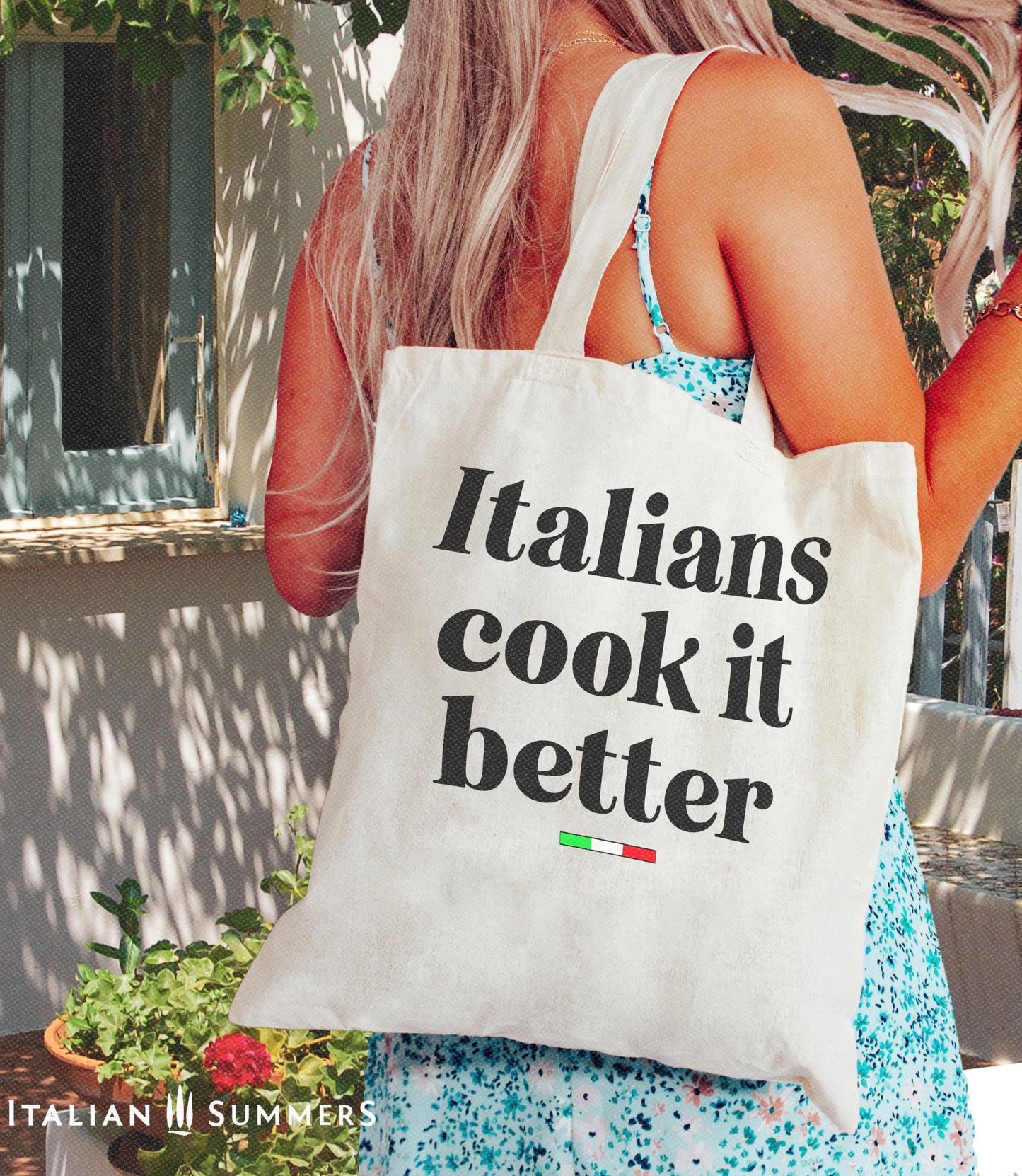 A Canvas tote bag with the quote Italians cook it better. Under the text in a black font there is a little bar with the happy colors of the Italian flag. in the background is a Mediterranean terrace with shady recesses and garden plants. Designed by Italian Summers