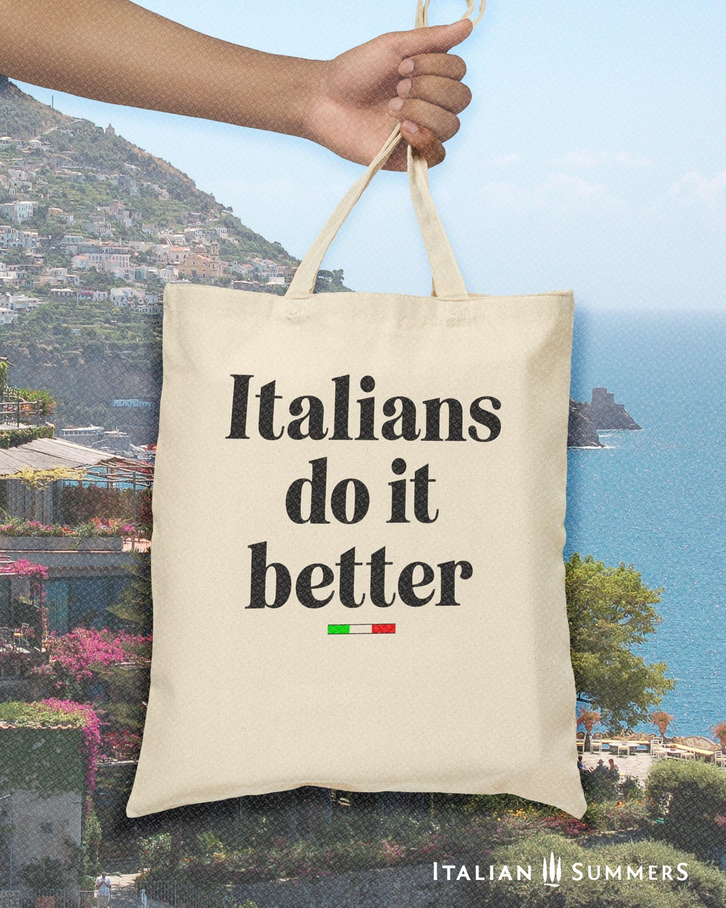 A Canvas tote bag with the quote Italians do it better. Under the text in a black font there is a little bar with the happy colors of the Italian flag. in the background is an Amalfi Coast landscape . Designed by Italian Summers