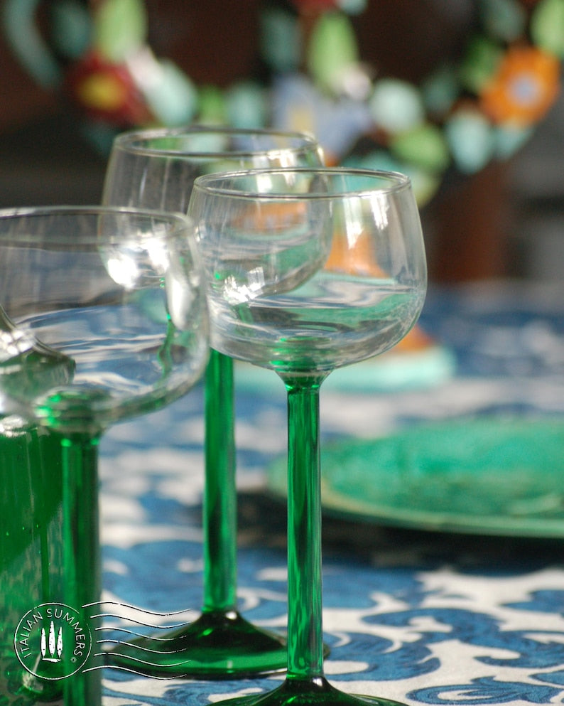An emerald green glass carafe stands surrounded by four green-stemmed wine glasses on top of a blue and white floral tablecloth with colorful ceramic plates with bright tomatoes in the background
