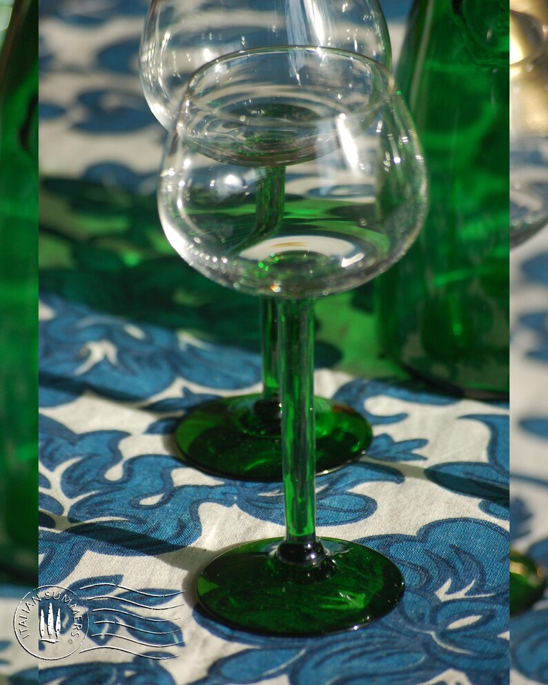 An emerald green glass carafe stands surrounded by four green-stemmed wine glasses on top of a blue and white floral tablecloth with colorful ceramic plates with bright tomatoes in the background