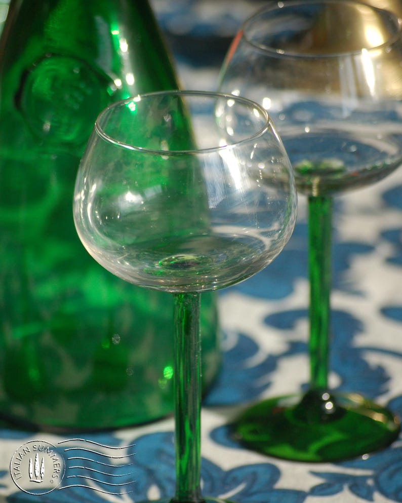 An emerald green glass carafe stands surrounded by four green-stemmed wine glasses on top of a blue and white floral tablecloth with colorful ceramic plates with bright tomatoes in the background