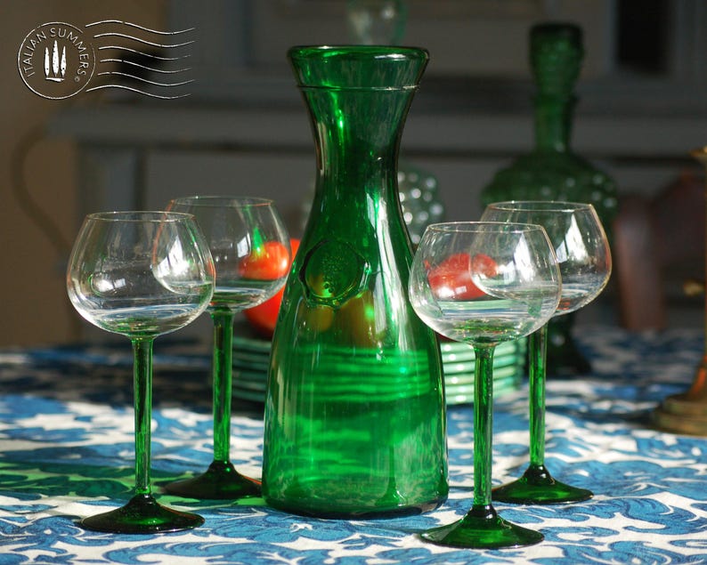An emerald green glass carafe stands surrounded by four green-stemmed wine glasses on top of a blue and white floral tablecloth with colorful ceramic plates with bright tomatoes in the background