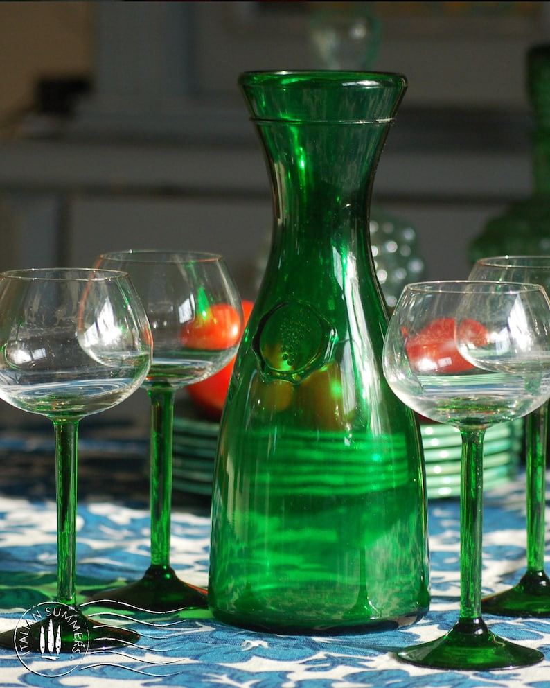 An emerald green glass carafe stands surrounded by four green-stemmed wine glasses on top of a blue and white floral tablecloth with colorful ceramic plates with bright tomatoes in the background
