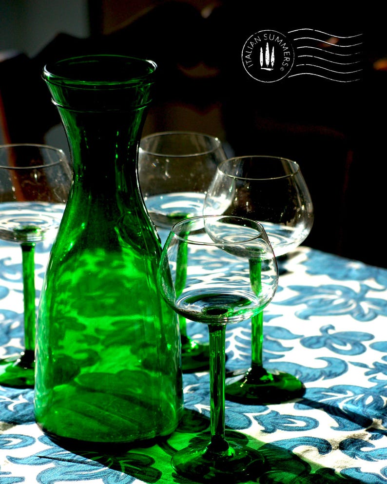 An emerald green glass carafe stands surrounded by four green-stemmed wine glasses on top of a blue and white floral tablecloth with colorful ceramic plates with bright tomatoes in the background