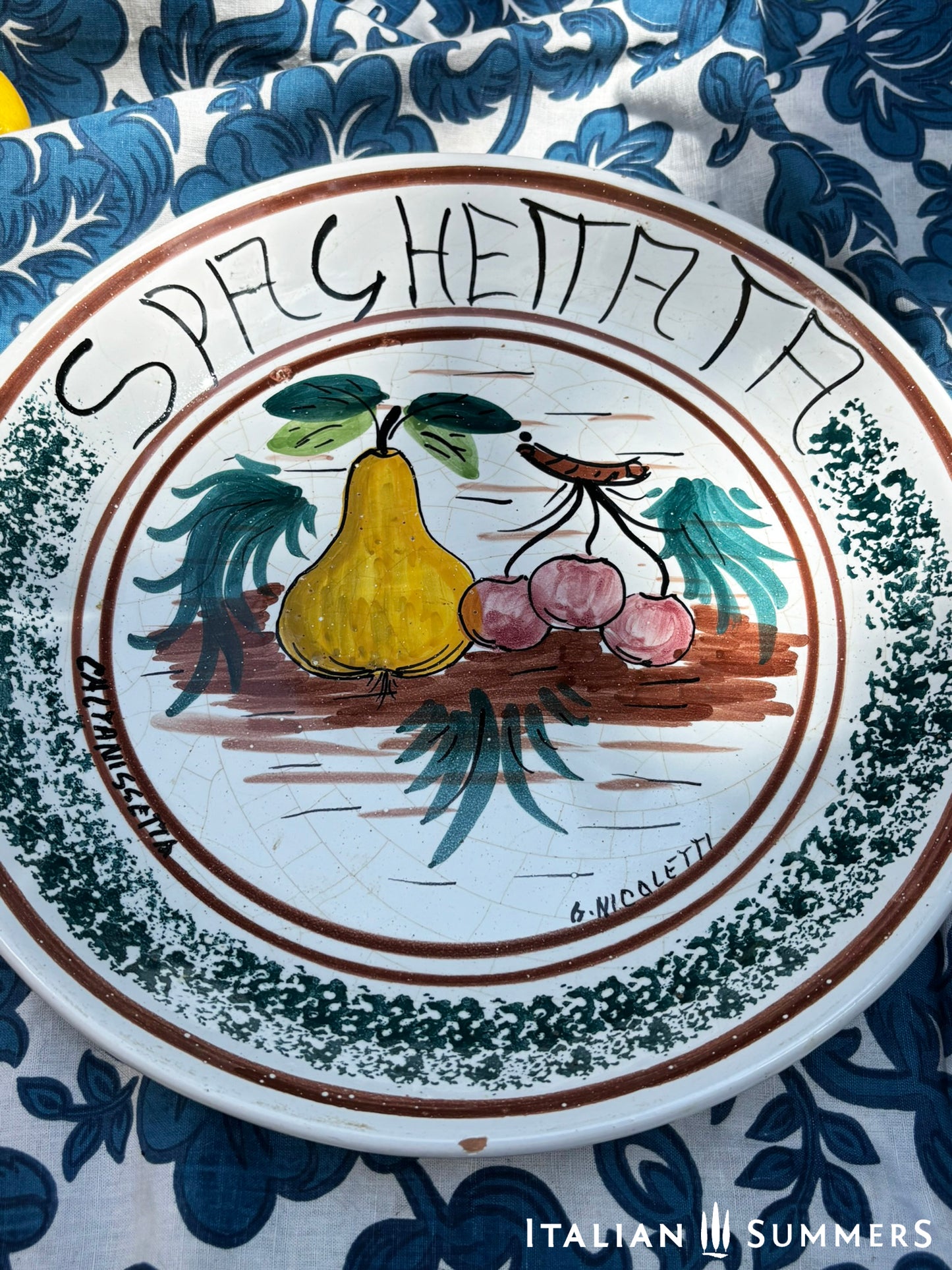 A large Sicilian vintage serving or Decorative plate with 'Spaghettata"' text and pear and cherries traditional Sicilian Maiolica illustration, on a blue floral patterned background.