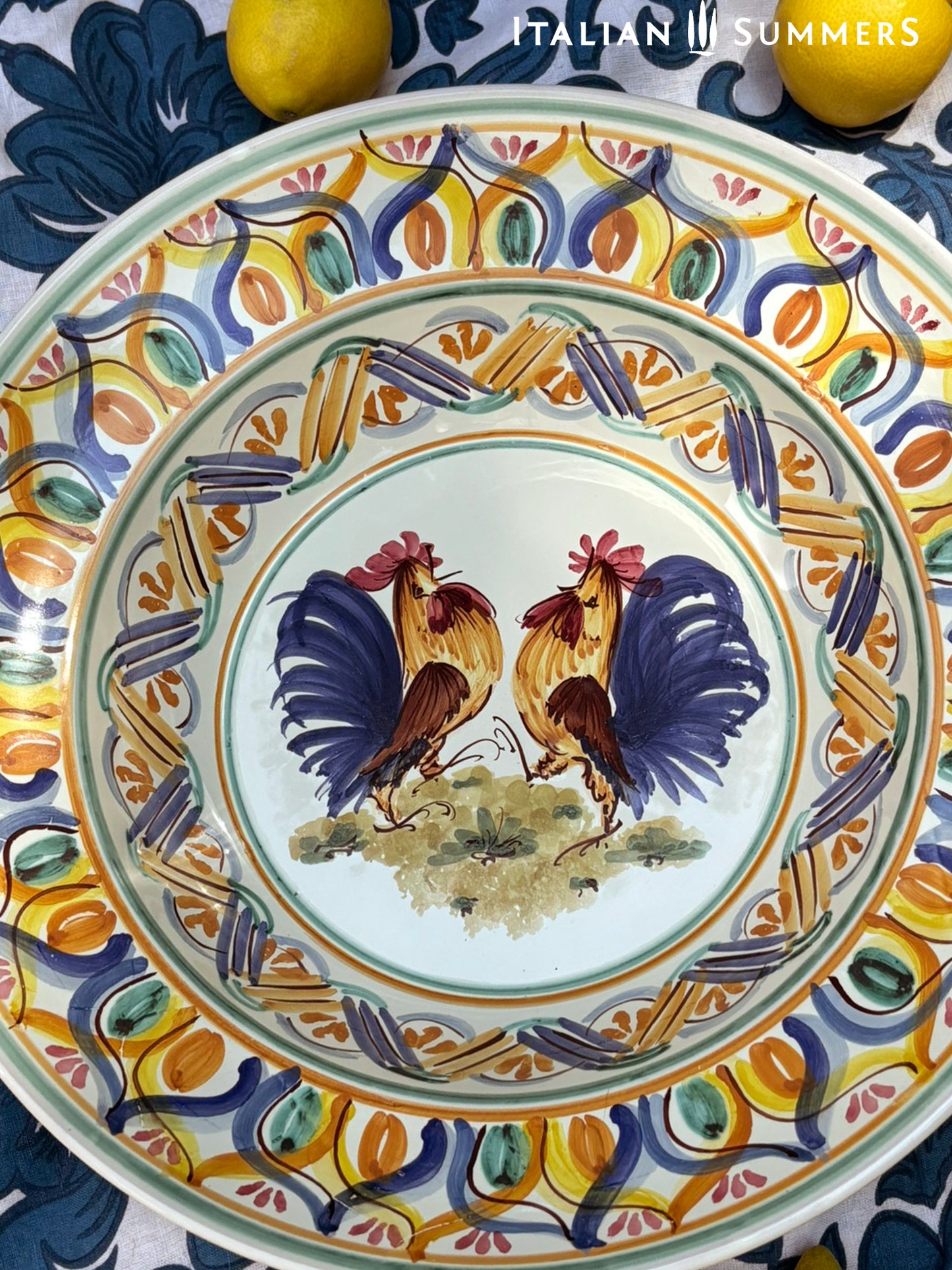 A large Apulian serving or decorative ceramic plate with rooster design on a blue patterned tablecloth with lemons.
