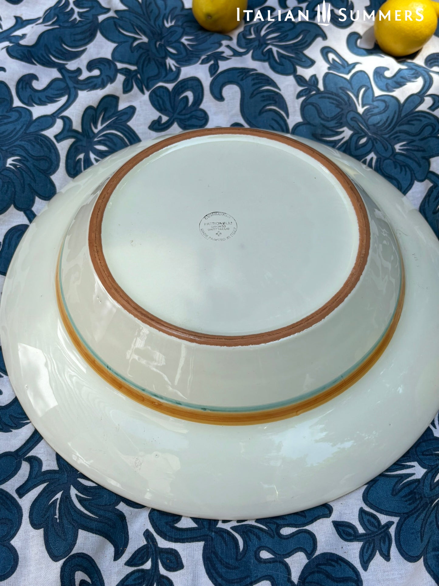 A large Apulian serving or decorative ceramic plate with rooster design on a blue patterned tablecloth with lemons.