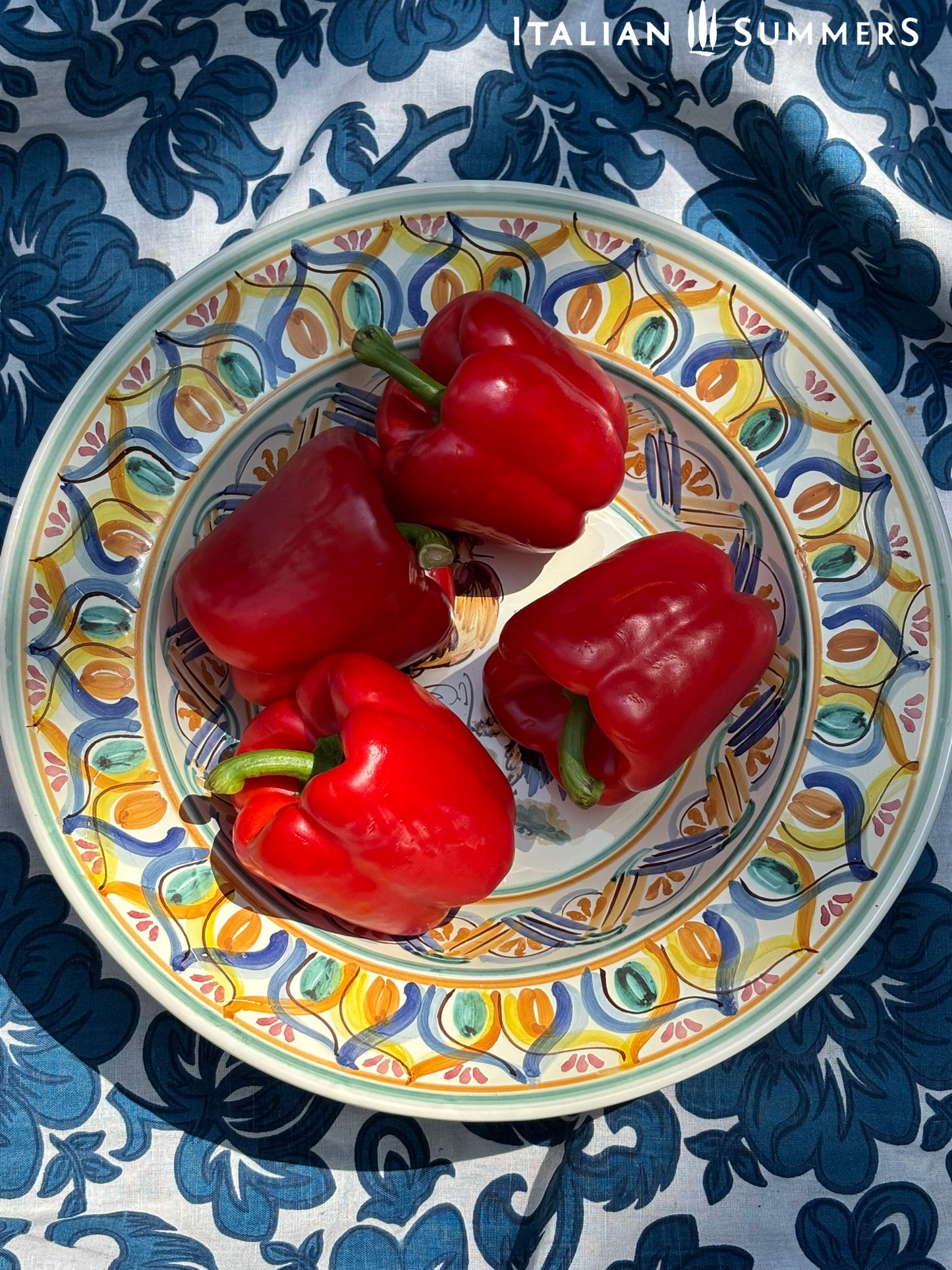 A large Apulian serving or decorative ceramic plate with rooster design on a blue patterned tablecloth with lemons.