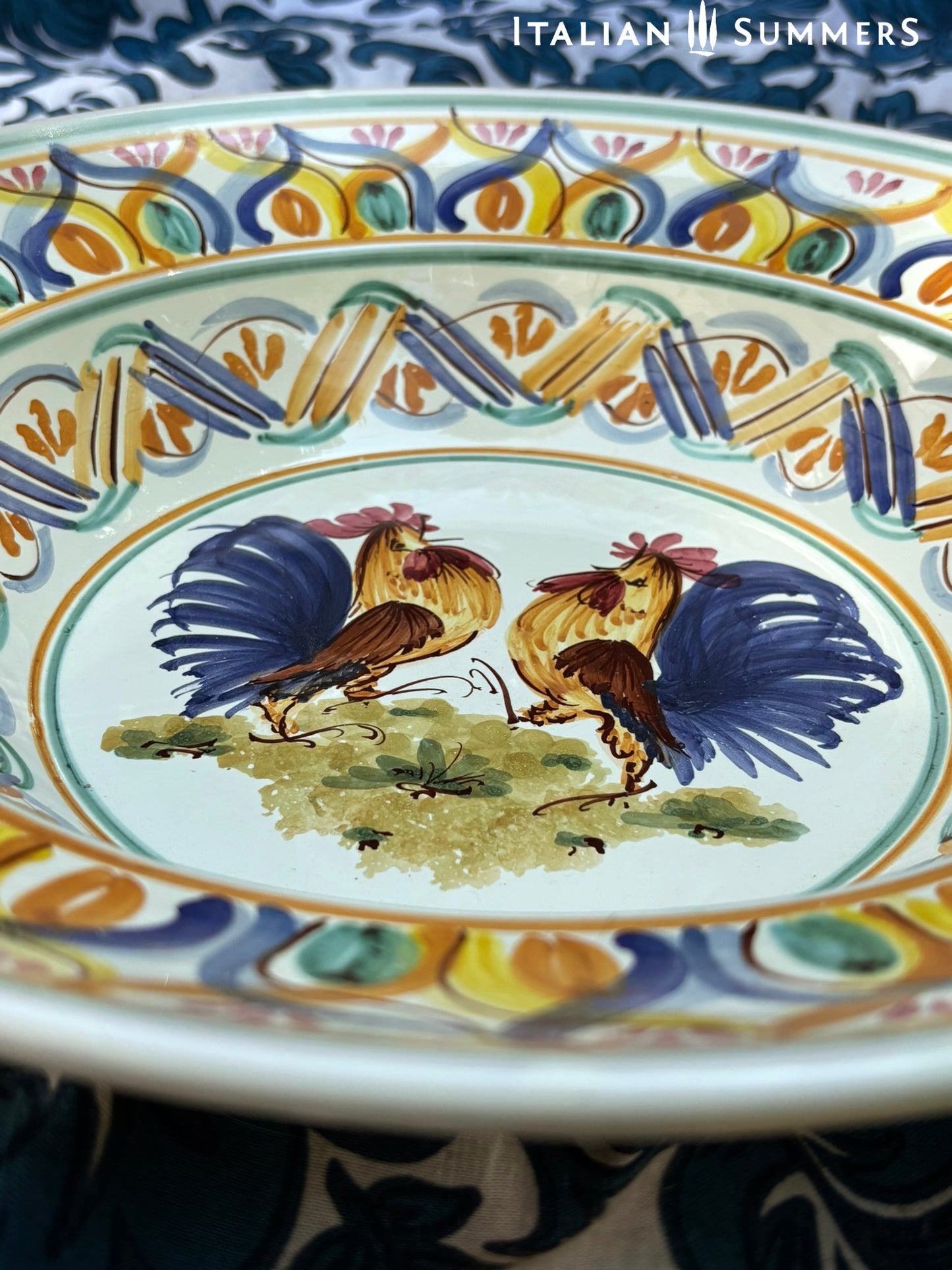 A large Apulian serving or decorative ceramic plate with rooster design on a blue patterned tablecloth with lemons.
