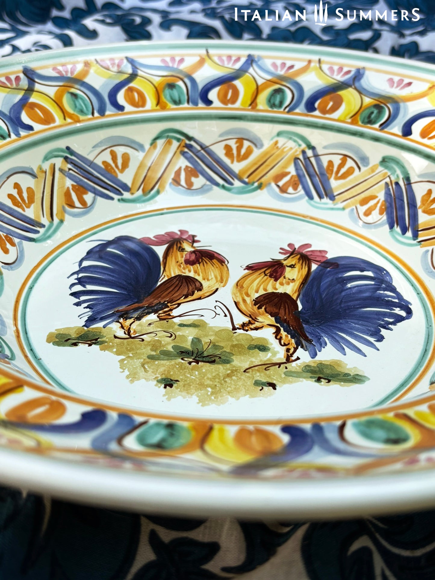 A large Apulian serving or decorative ceramic plate with rooster design on a blue patterned tablecloth with lemons.