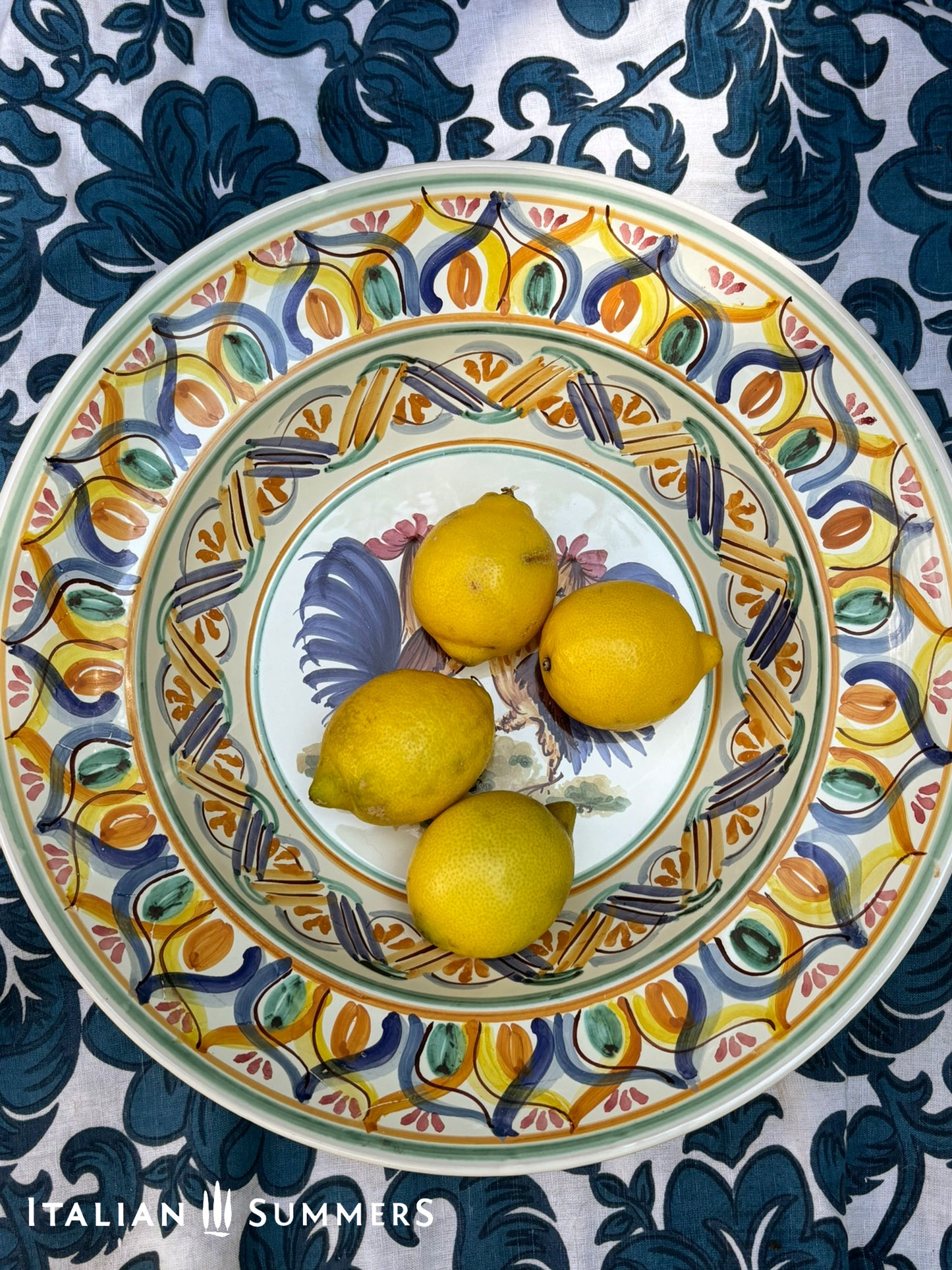 A large Apulian serving or decorative ceramic plate with rooster design on a blue patterned tablecloth with lemons.