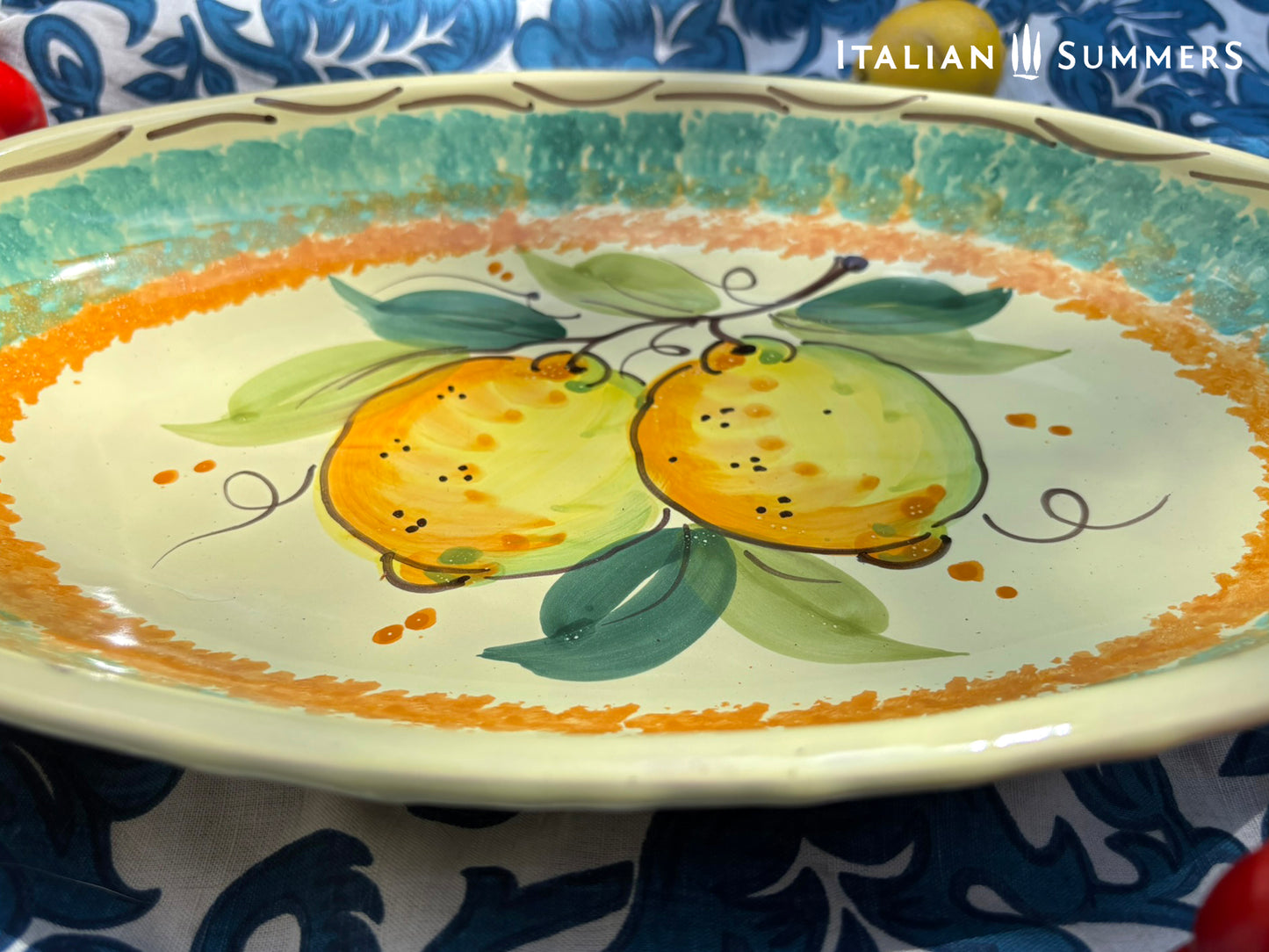 A large oval serving or Decorative Sicilian ceramic plate with alemon design on a blue floral tablecloth, featuring traditional maiolica enamel colors in the 'fangotto' banding style reminiscent of baroque Sicily.