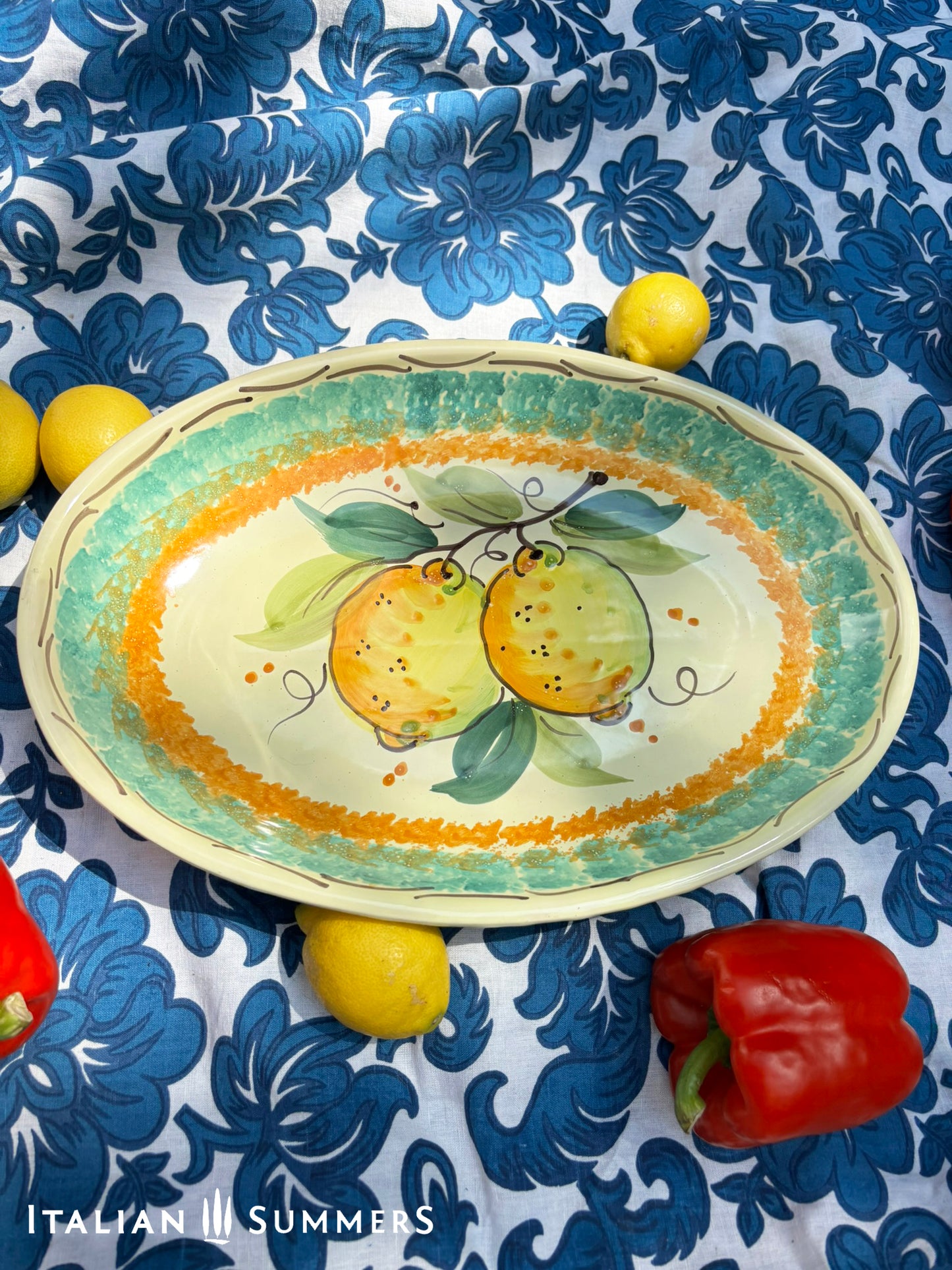 A large oval serving or Decorative Sicilian ceramic plate with alemon design on a blue floral tablecloth, featuring traditional maiolica enamel colors in the 'fangotto' banding style reminiscent of baroque Sicily.