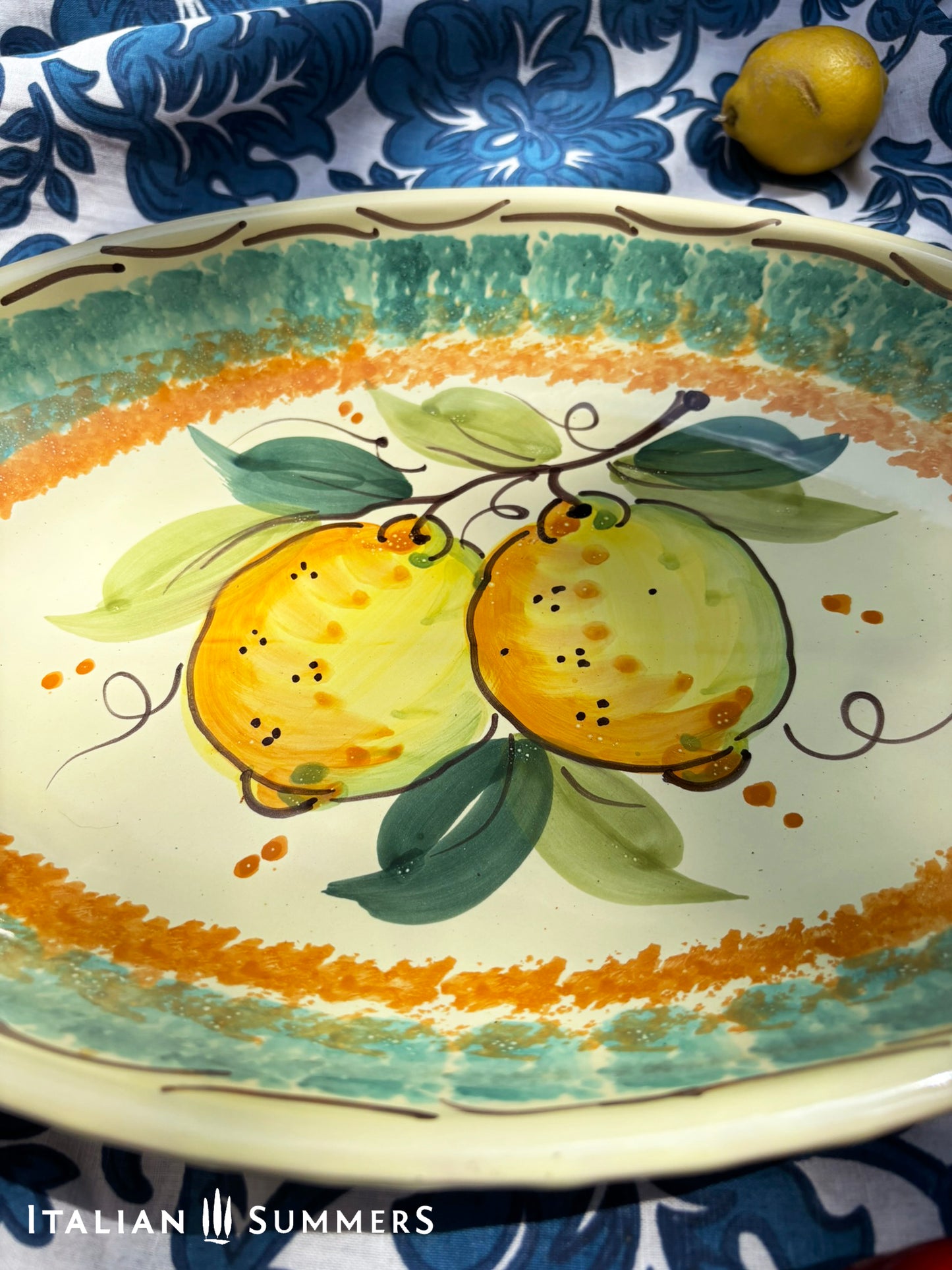 A large oval serving or Decorative Sicilian ceramic plate with alemon design on a blue floral tablecloth, featuring traditional maiolica enamel colors in the 'fangotto' banding style reminiscent of baroque Sicily.