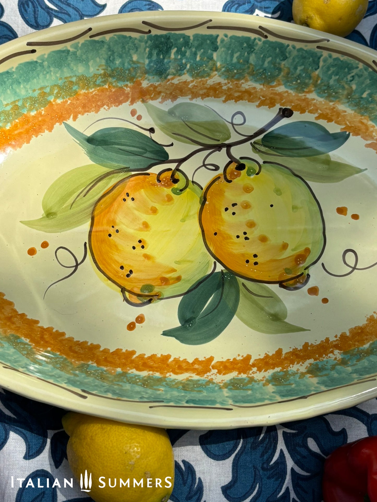 A large oval serving or Decorative Sicilian ceramic plate with alemon design on a blue floral tablecloth, featuring traditional maiolica enamel colors in the 'fangotto' banding style reminiscent of baroque Sicily.