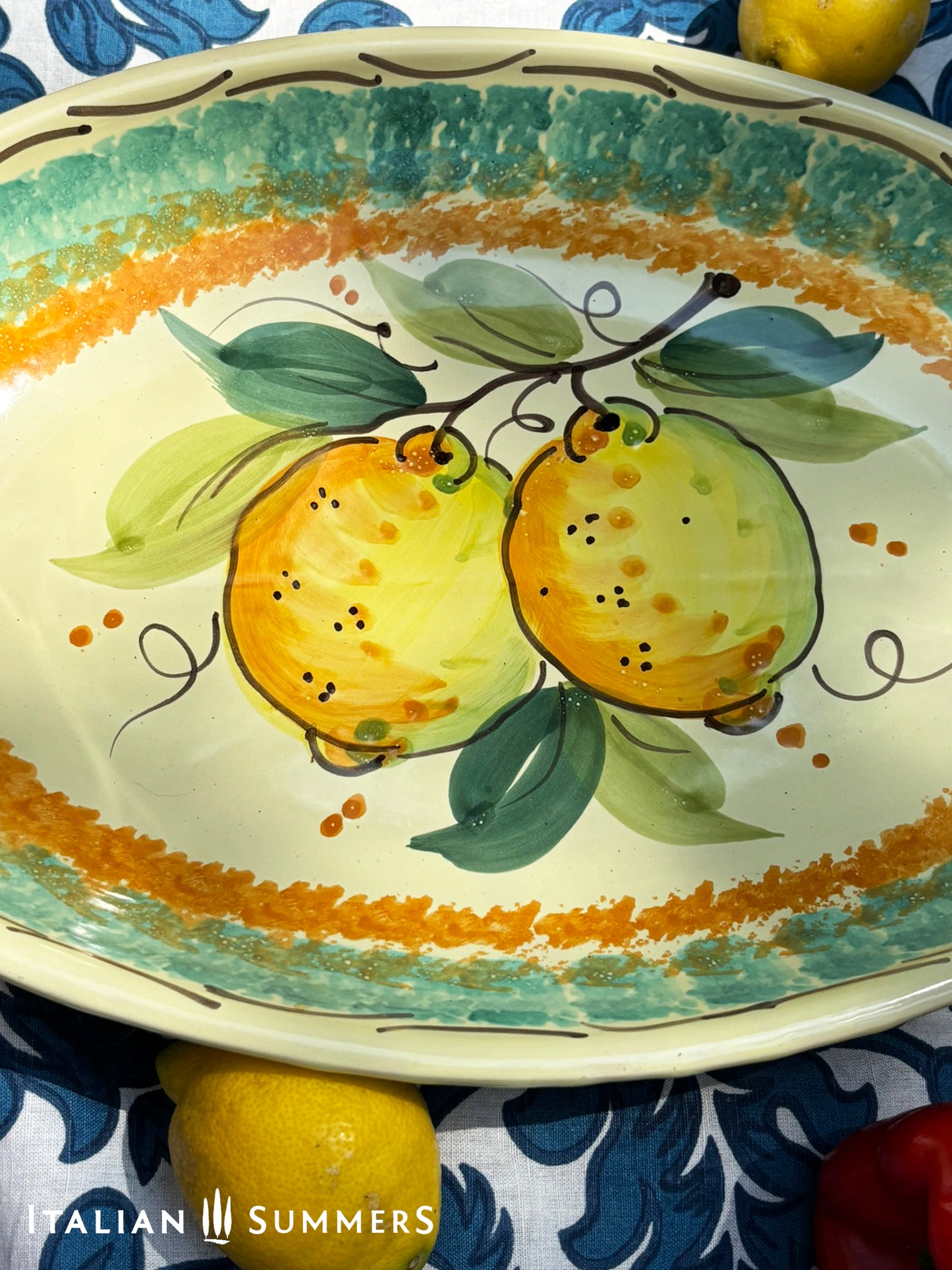 A large oval serving or Decorative Sicilian ceramic plate with alemon design on a blue floral tablecloth, featuring traditional maiolica enamel colors in the 'fangotto' banding style reminiscent of baroque Sicily.