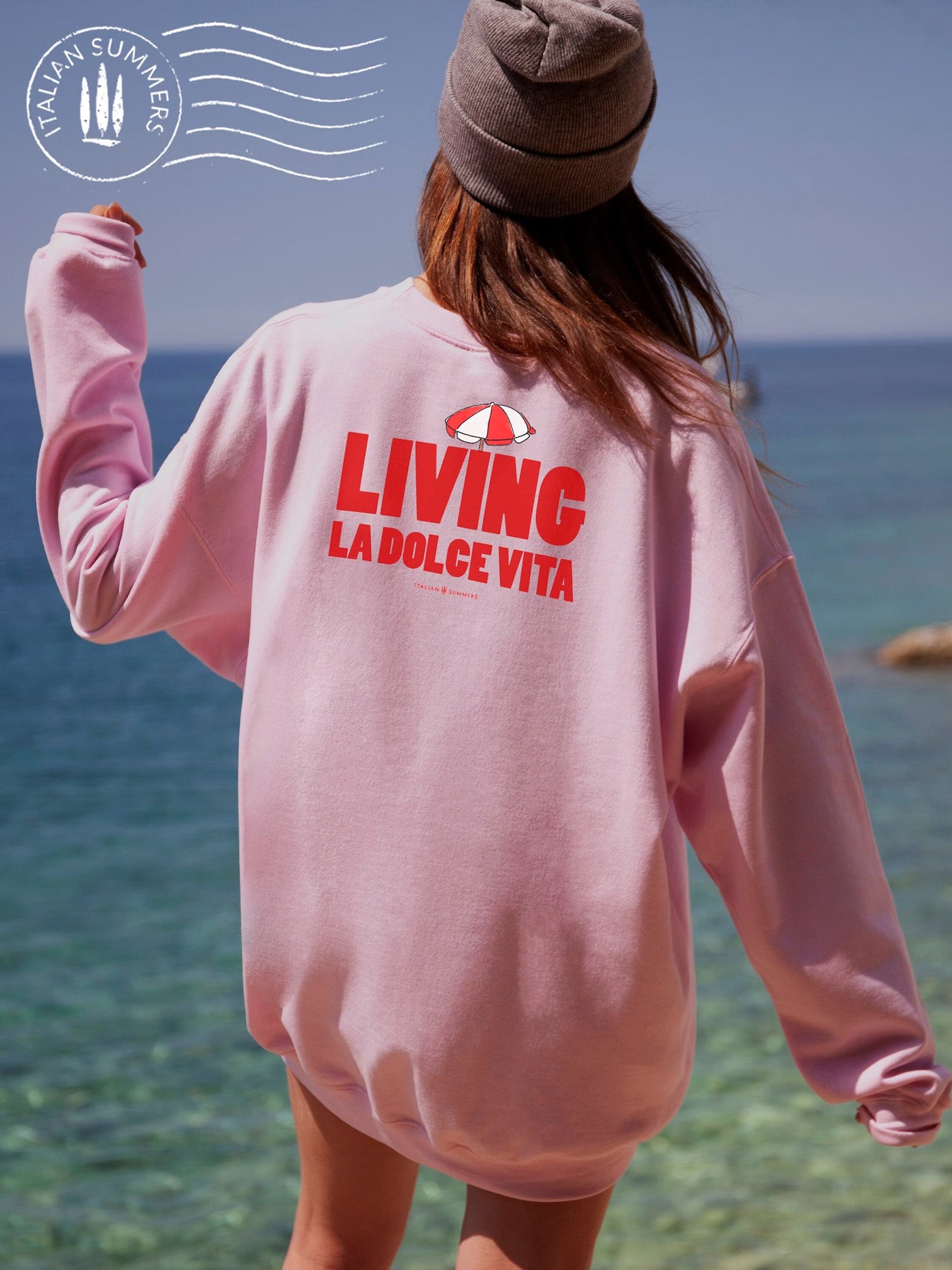 Soft and comfortable Italy-inspired  pink sweatshirt is embroidered in the front with the text 'Living la dolce Vita' in red bold letters and a small red and white beach umbrella on top. It features a large full-size print in the back with the same text and the same red and white beach umbrella.