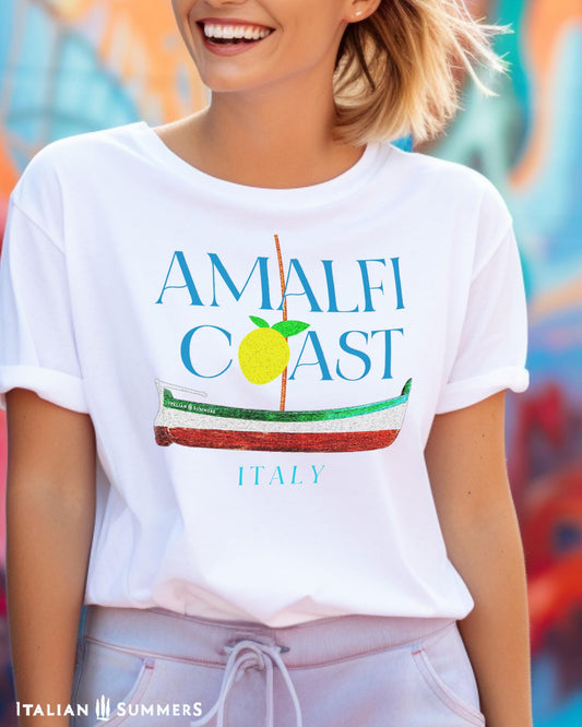 A smiling blonde woman wearing a white T shirt with blue text: AMALFI COAST, the O in Coast is a yellow stylized lemon with two leaves.
Under the text is a traditional Amalfi coast boat called a Gozzo, in the traditional colors of the Italian flag, green, white, red.