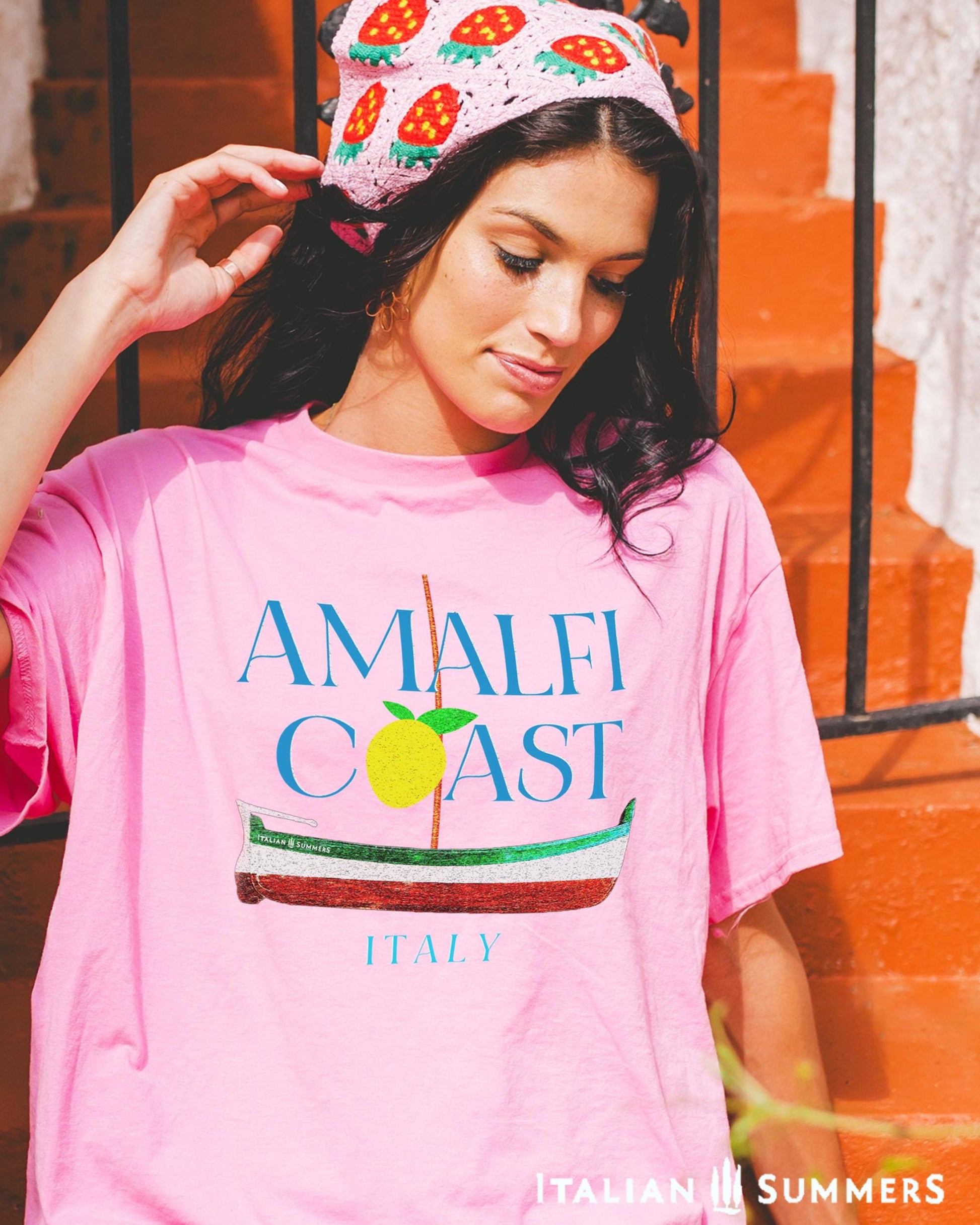 A smiling brunette woman wearing a pink T shirt with blue text: AMALFI COAST, the O in Coast is a yellow stylized lemon with two leaves.
Under the text is a traditional Amalfi coast boat called a Gozzo, in the traditional colors of the Italian flag, green, white, red.
The word ITALY in light blue end the composition on the bottom.