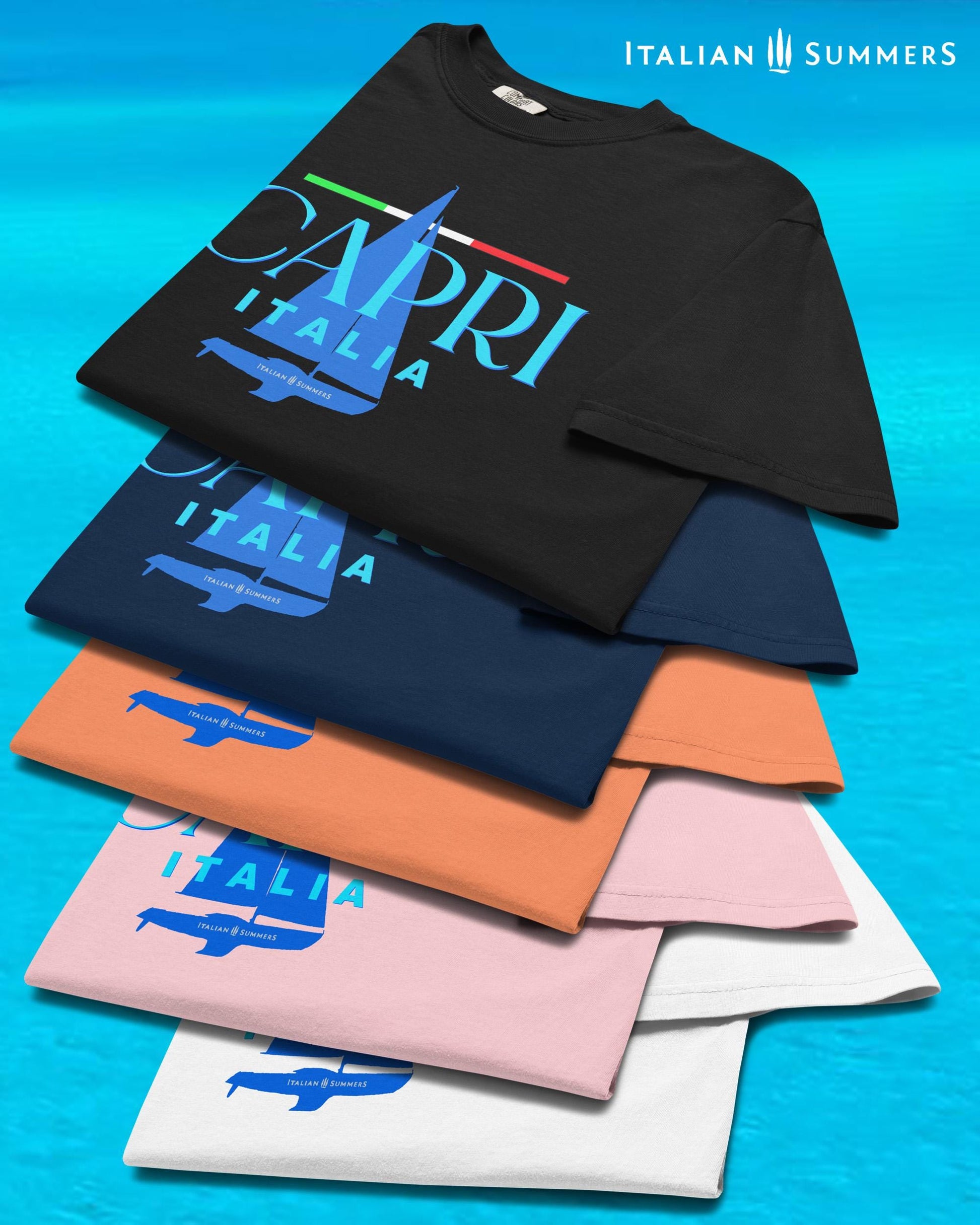A stack of different color T shirts with a frontal print featuring a marine blue silhouette of a sailboat and a large text: CAPRI, ITALIA, with a thin Italian flag as a chevron, looking like a sailing shirt for sportsmen or Mediterranean Sea enthusiasts.
