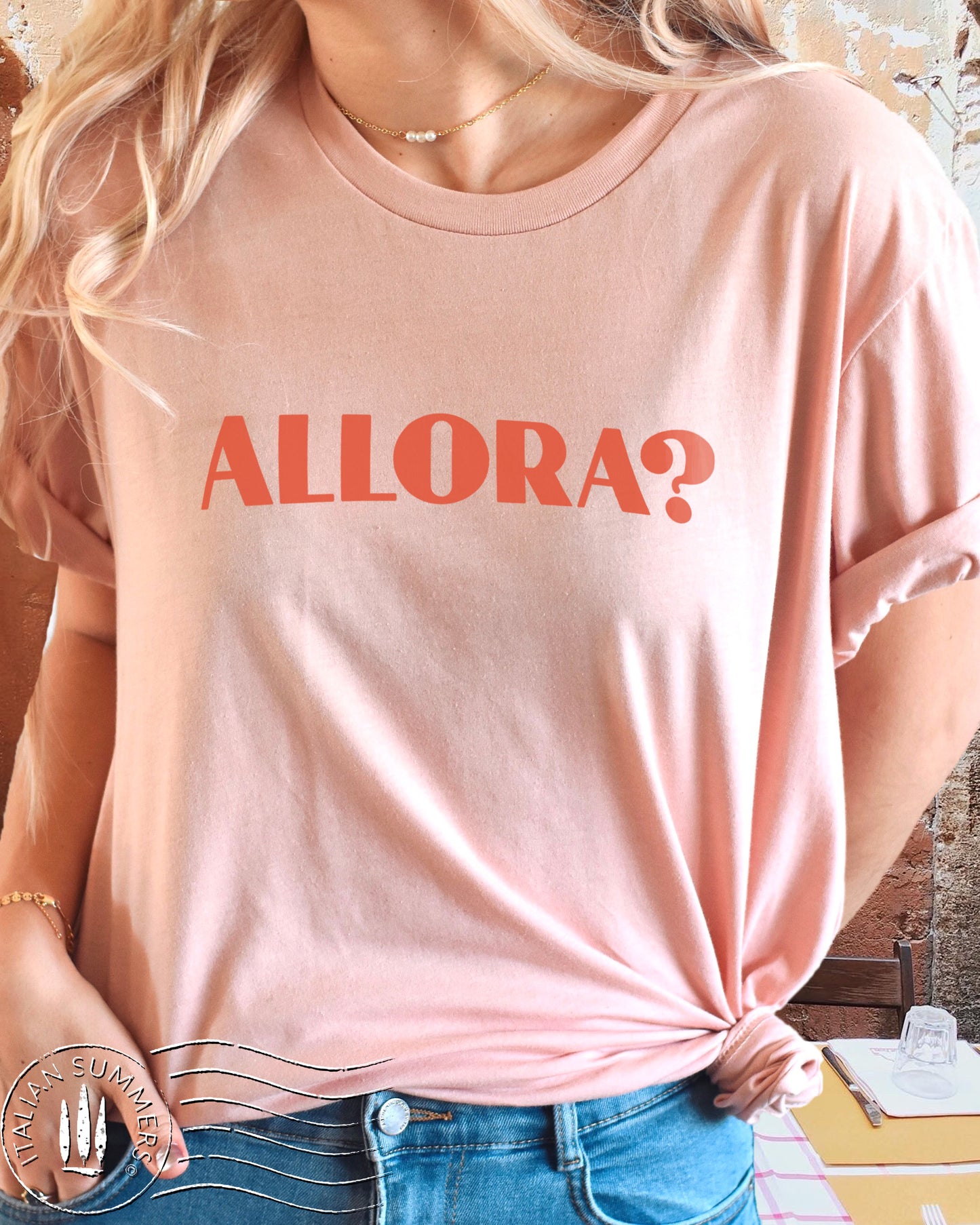 A girl wearing a pink Italy inspired t-shirt with red text of the Italian expression: Allora?, Used all the time in Italian language. The color of the shirt is a soft pastel peach with the Allora expression in terra. Designed and sold by Italian Summers