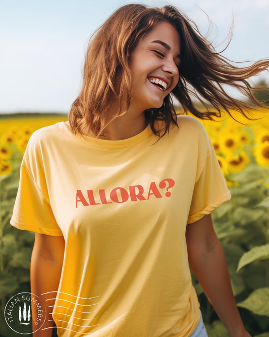 A girl stands in a field of Italian sunflowers. She wears a yellow Italy inspired t-shirt with a red text  of the Italian expression: Allora?, Used all the time in Italian language. The color of the shirt is a soft pastel peach with the Allora expression in terra. Designed and sold by Italian Summers
