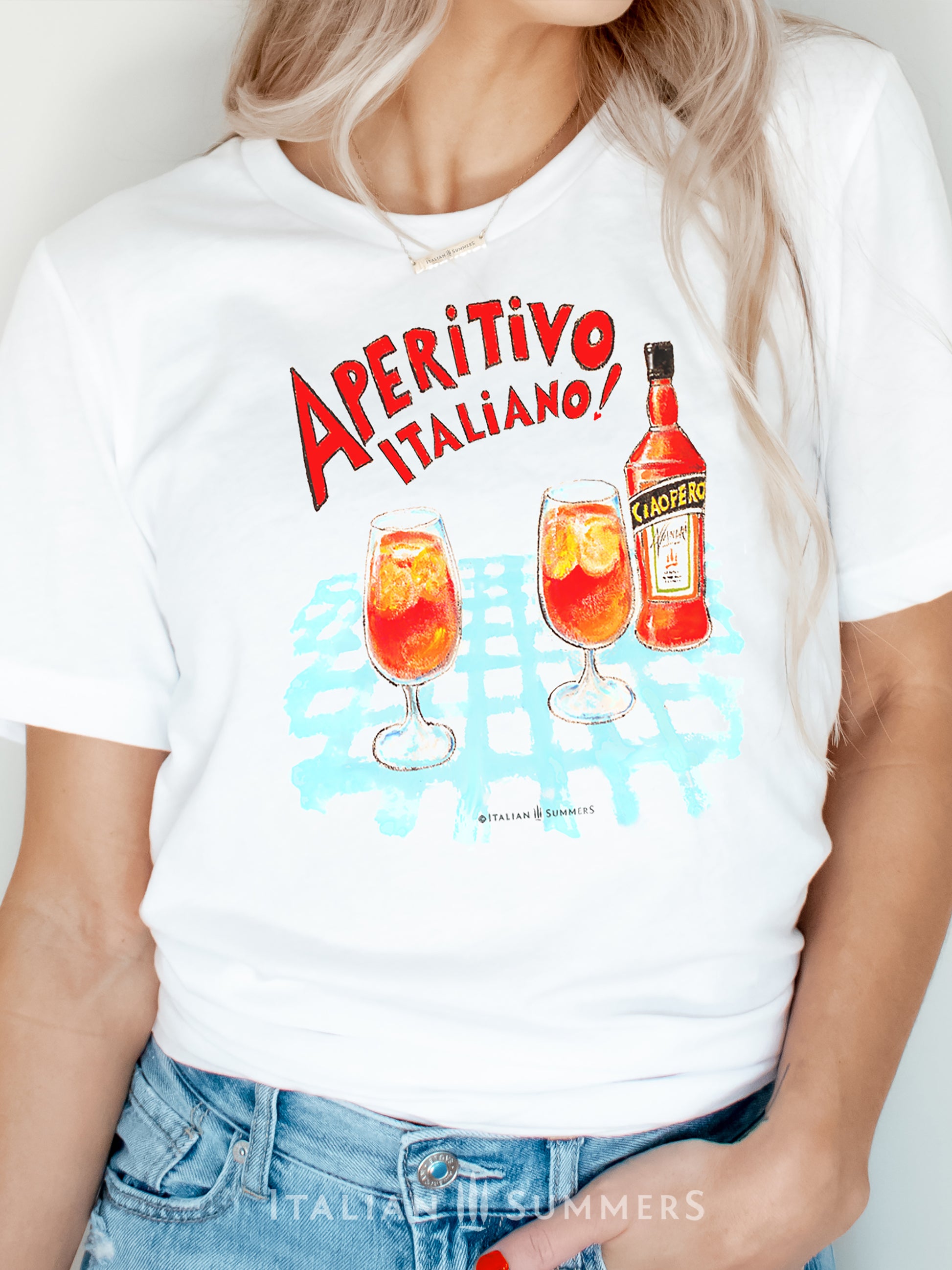 Italy inspired white unisex cotton T shirt with a print of a watercolor illustration of a bottle of red Italian aperritif and two spritz glasses filled with  ice, orange slices and spritz. A large red hand-painted text states: " Aperitivo Italiano"