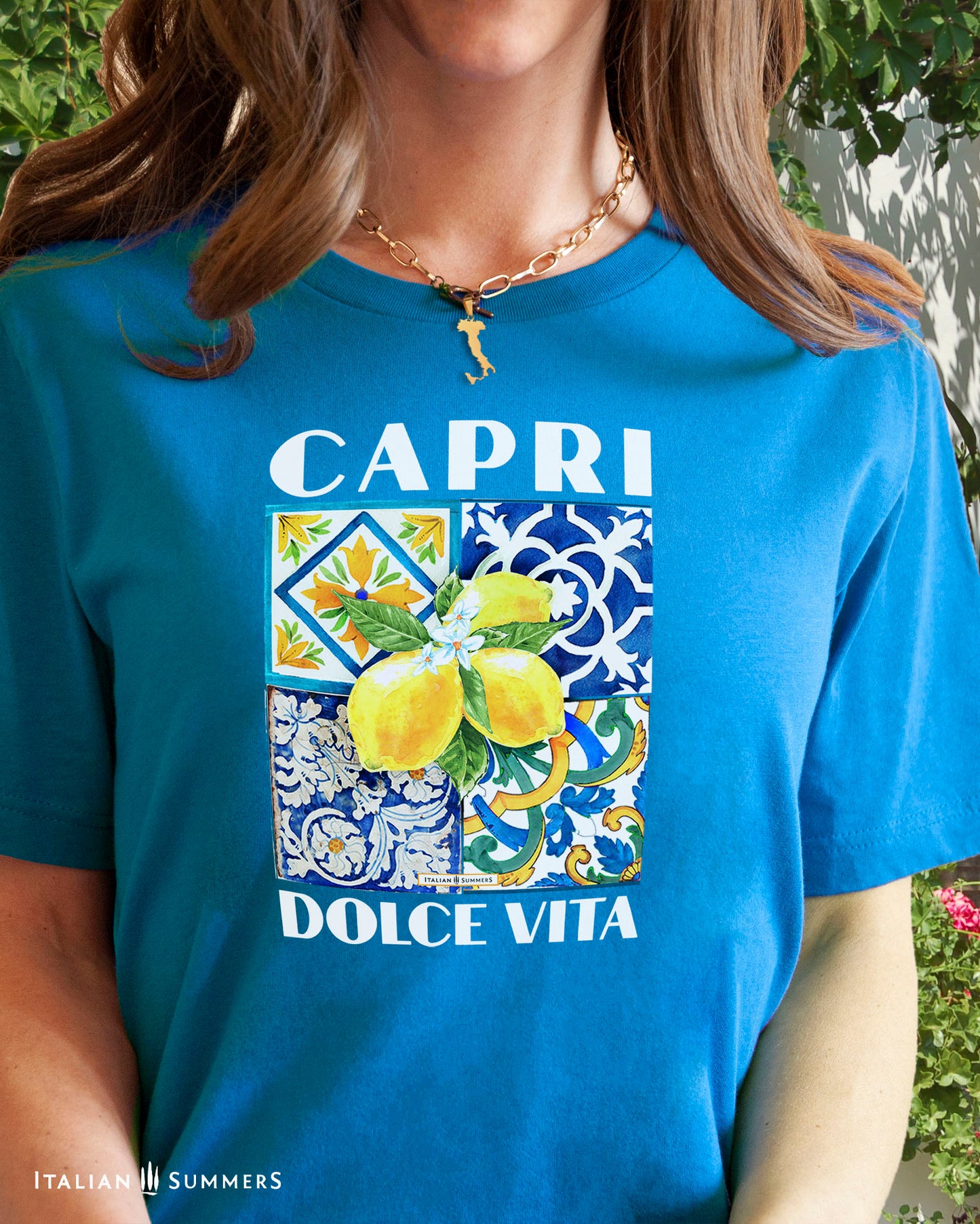 An all cotton T shirt with a Mediterranean tile design on the chest and a large cluster of Amalfi Coast lemons on top of the tiles. The lemons are bright yellow with green leaves and flowers. A blue text states: CAPRI DOLCE VITA