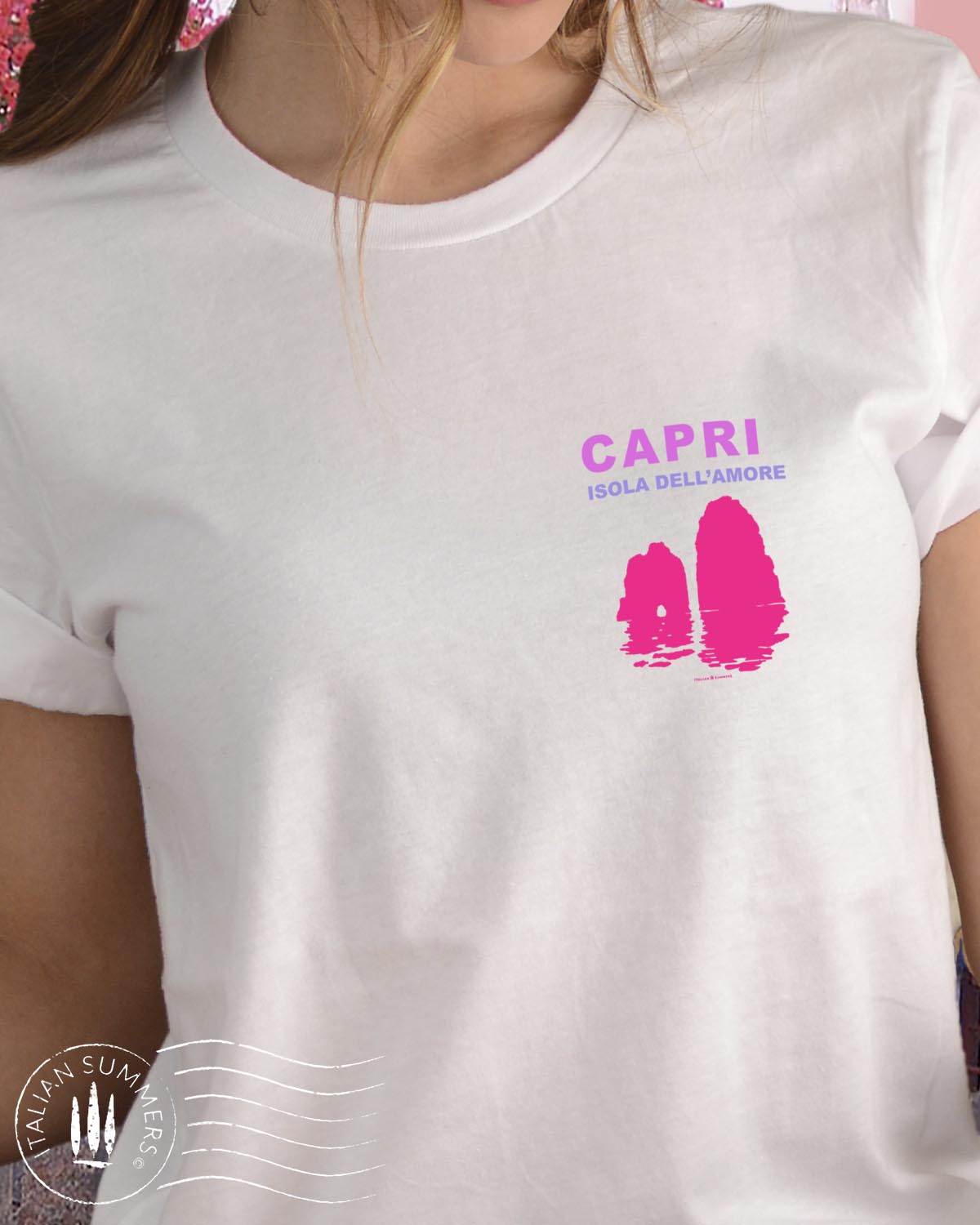 A youg woman stands in front of a quaint Italian island restaurant covered in pink bougainvilleas. She is standing with her back to us wearing a soft white cotton shirt with a pink and fuchsia print depicting the Faraglioni islets of the famous island of Capri. The text on the print states " Capri Isola dell'Amore" Capri, island of love.